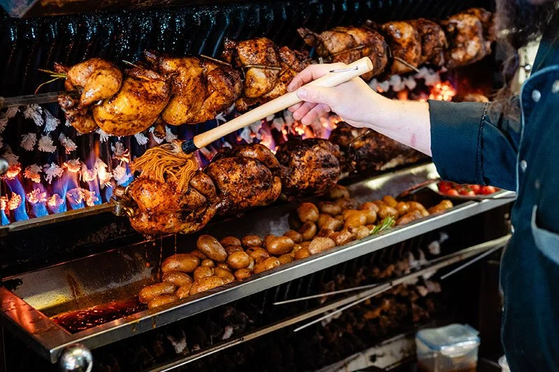 Person, die Hähnchen am Spieß über offener Flamme grillt, mit Kartoffeln und Beilagen im Hintergrund.