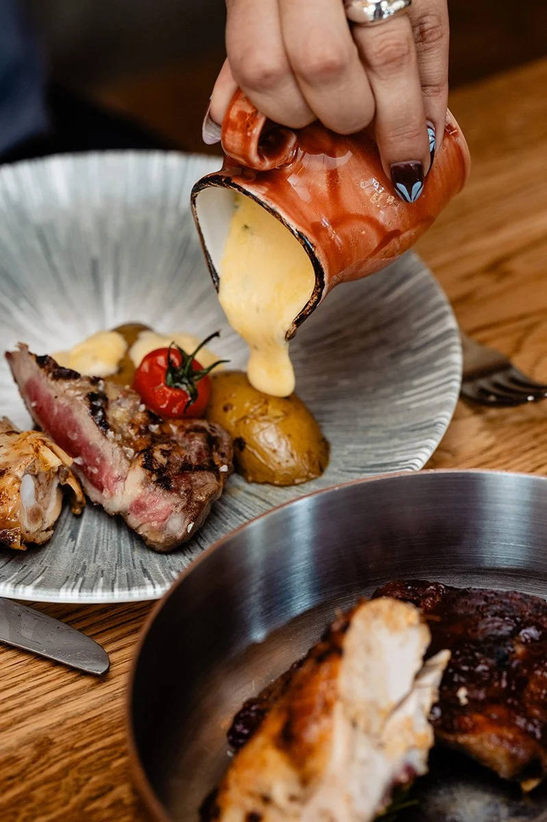 Jemand gießt Sauce über ein Steak mit Kartoffeln auf einem Teller, daneben liegt eine kleine Tomate. Im Vordergrund ist eine weitere Platte mit Fleisch.