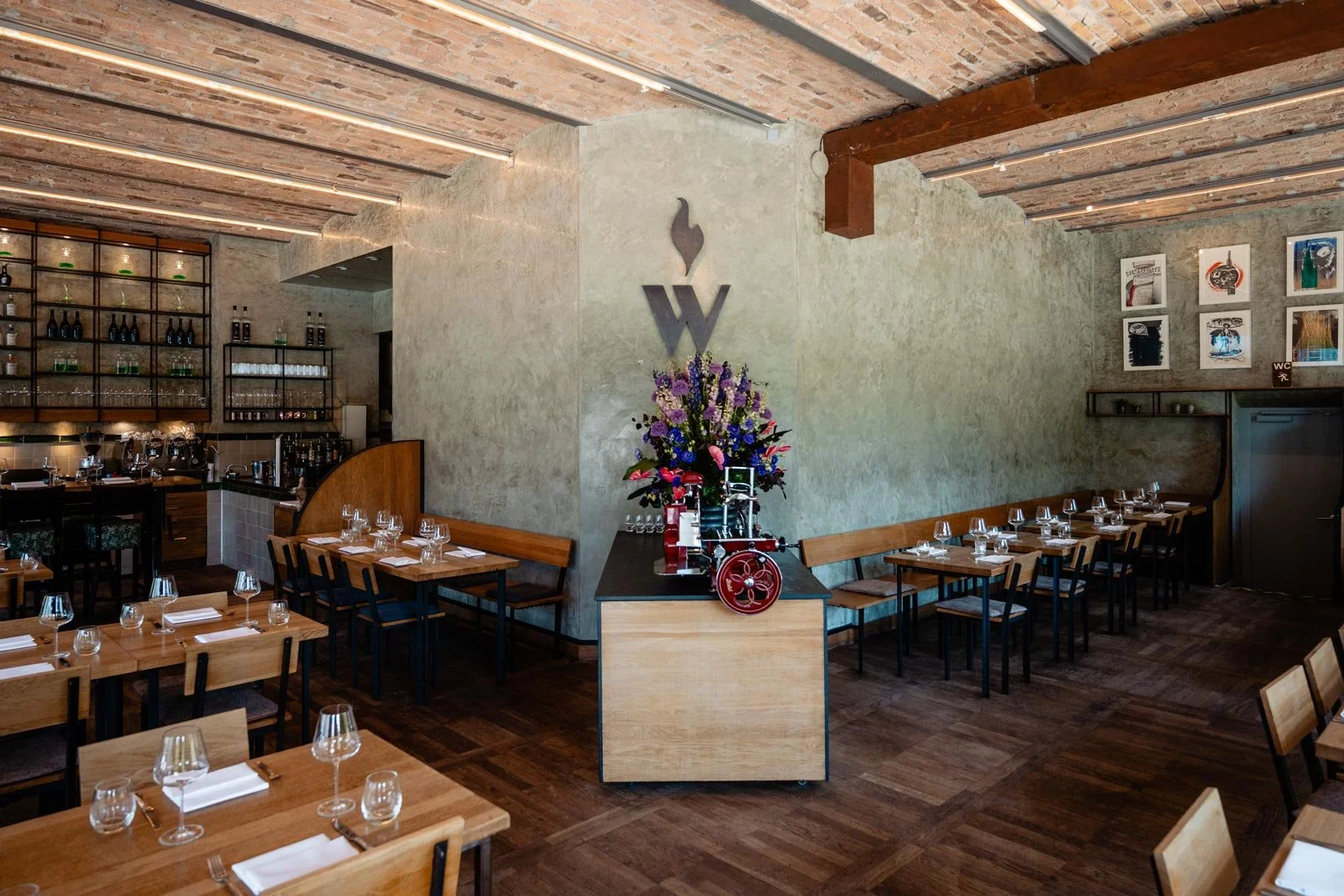 Innenraum der Rotisserie Weingrün mit Tischen, Weingläsern und Besteck, einer Blumenarrangement auf einem Podest, Holzfußboden, erleuchteter Decke und Kunstwerken an den Wänden.