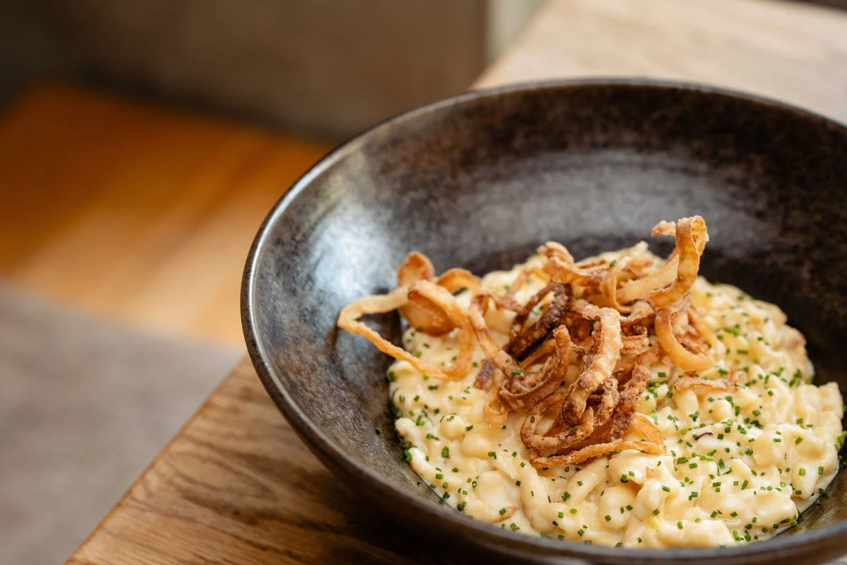 Ein dunkler Teller mit Käsespätzle und knusprigen Zwiebelringen.