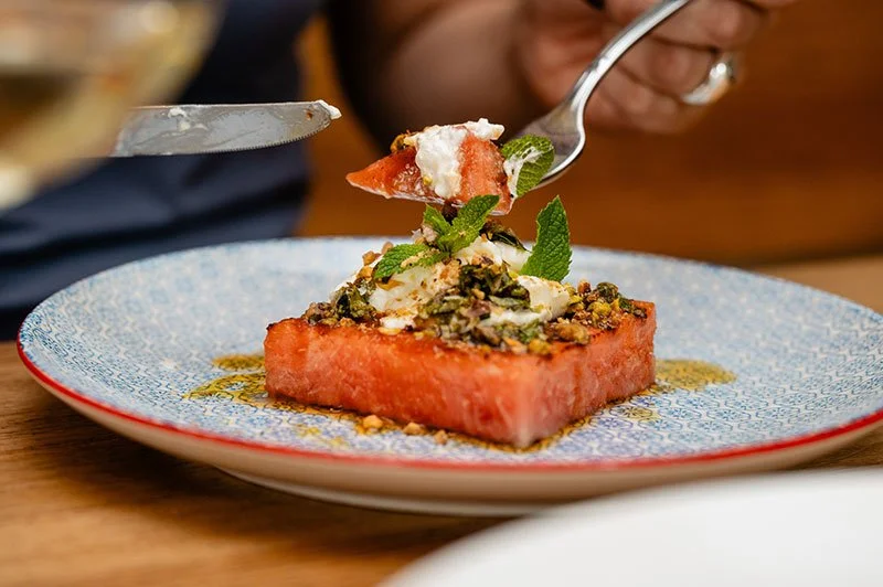 Ein Dessert mit einer großen Wassermelonenquadrat, Joghurt, Minzblättern und Pistazien auf einem gedeckten Tisch.