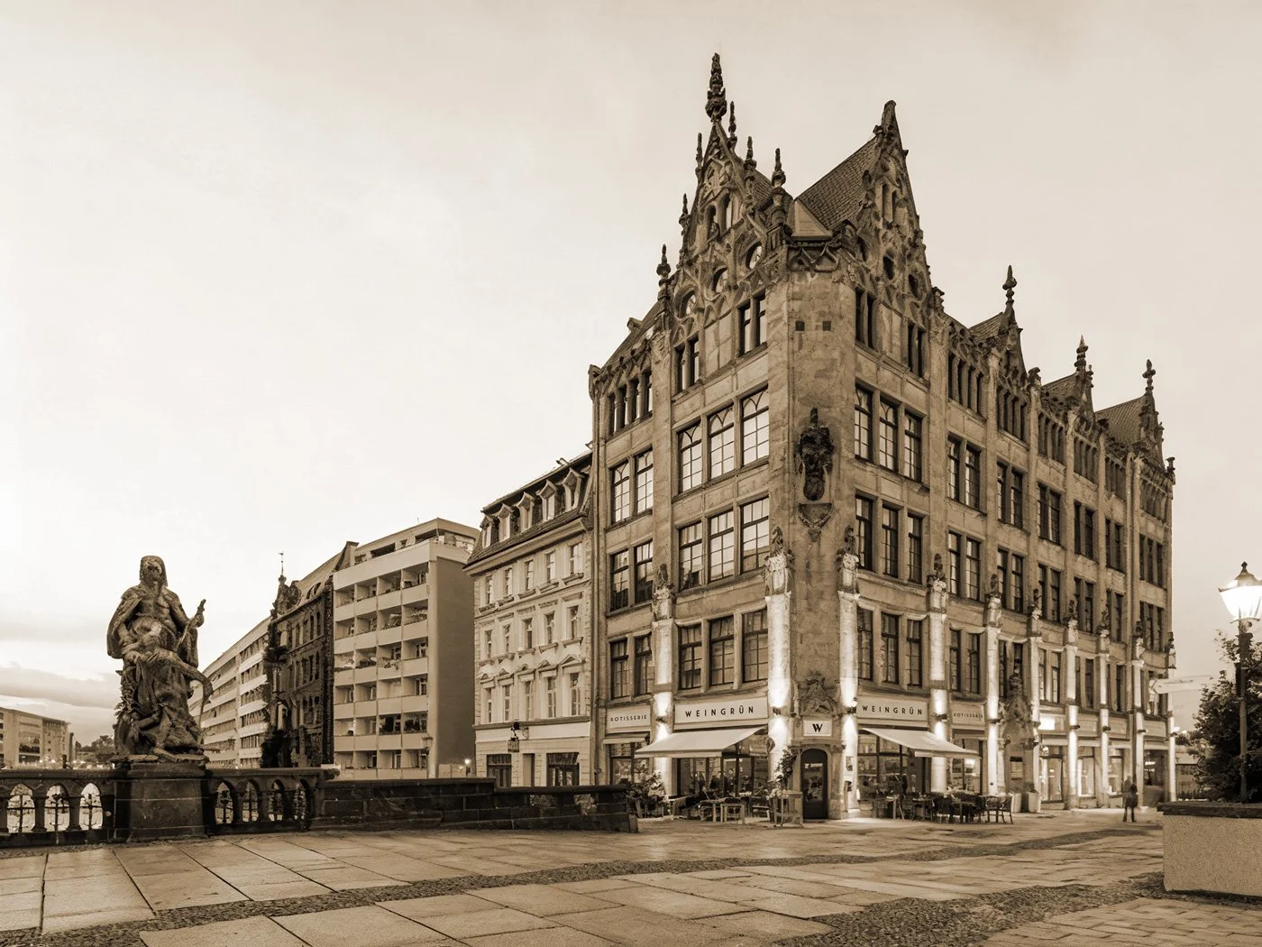 Historisches Gebäude mit gotischer Architektur, umgeben von modernen Gebäuden und Skulptur an der Flusspromenade in sepia Ton, abends, Überdachte Terrassen der Rotisserie Weingrün.