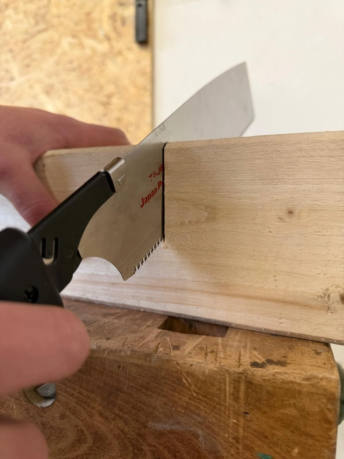 Une main tient une scie japonaise pour couper un morceau de bois clair fixé sur une surface en bois plus foncé.