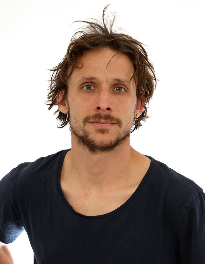 Un homme avec des cheveux en bataille, une barbe légère, portant un t-shirt noir, face à un fond blanc.