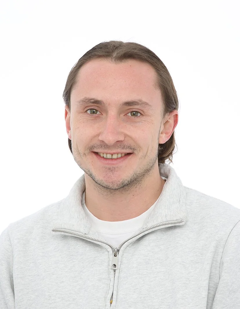 Un homme souriant avec des cheveux bruns, portant un sweat gris à col zippé, sur fond blanc.