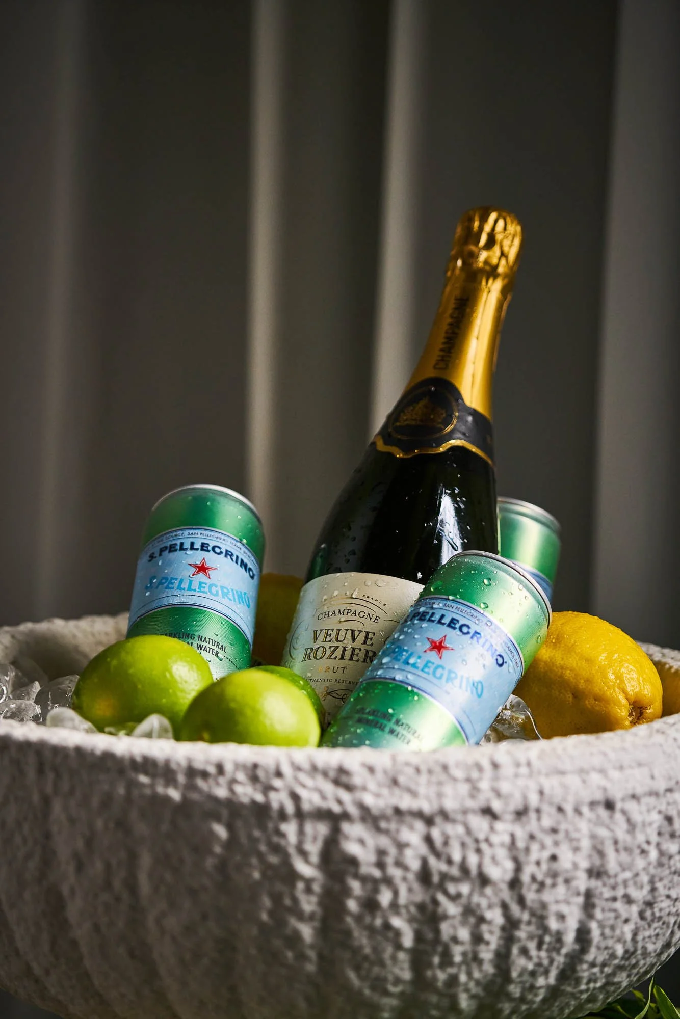 A large bowl filled with ice, containing a bottle of Veuve Clicquot champagne, two cans of San Pellegrino sparkling water, two green limes, and a yellow lemon.