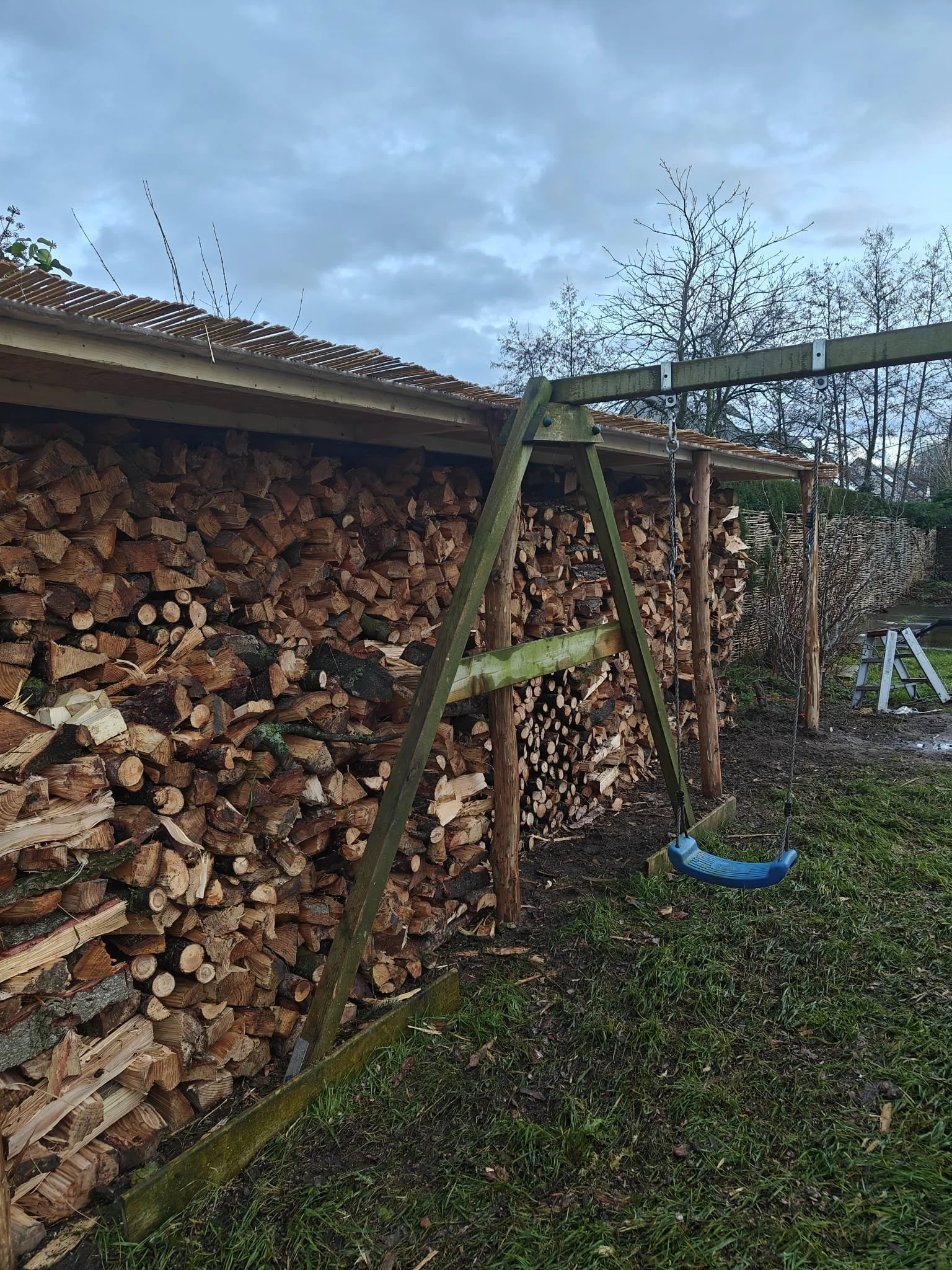 Een schommel in een tuin met opgestapeld hout op de achtergrond.