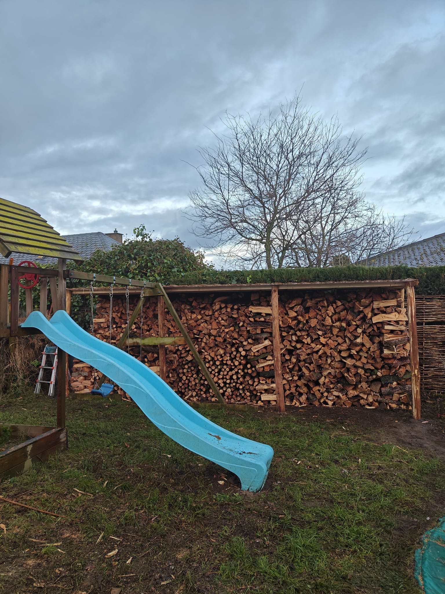 Een blauwe glijbaan in een tuin met stapel hout op de achtergrond.