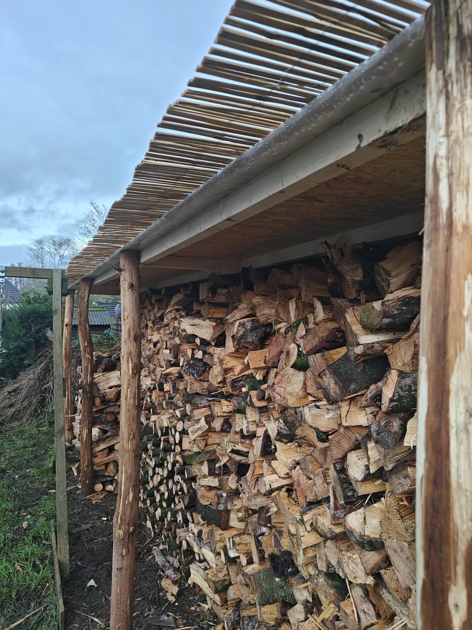 Een houtopslag met gestapelde houtblokken onder een afdak van houten planken.