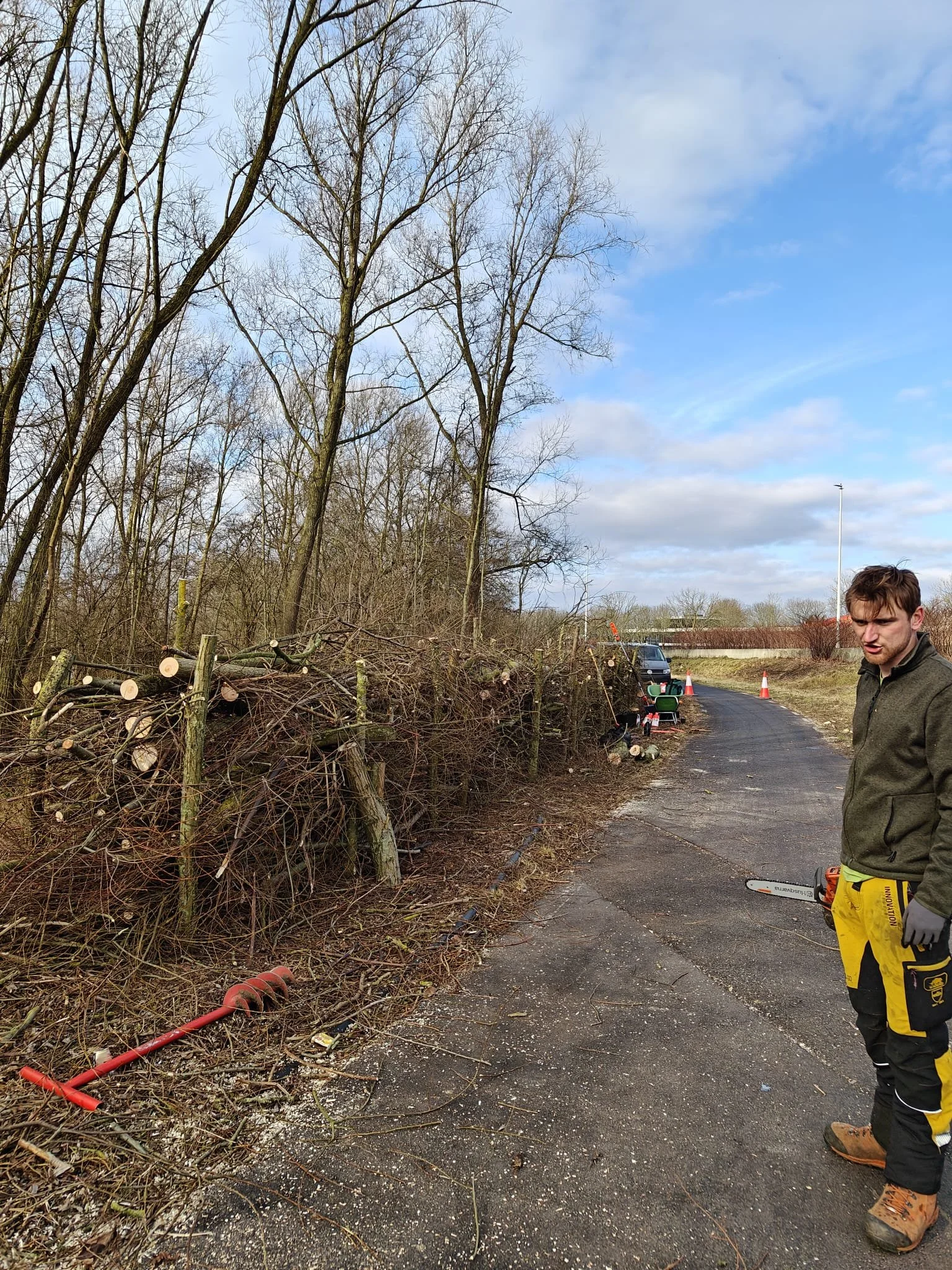 Man met een kettingzaag staat op een weg naast afgezaagde bomen en takken.