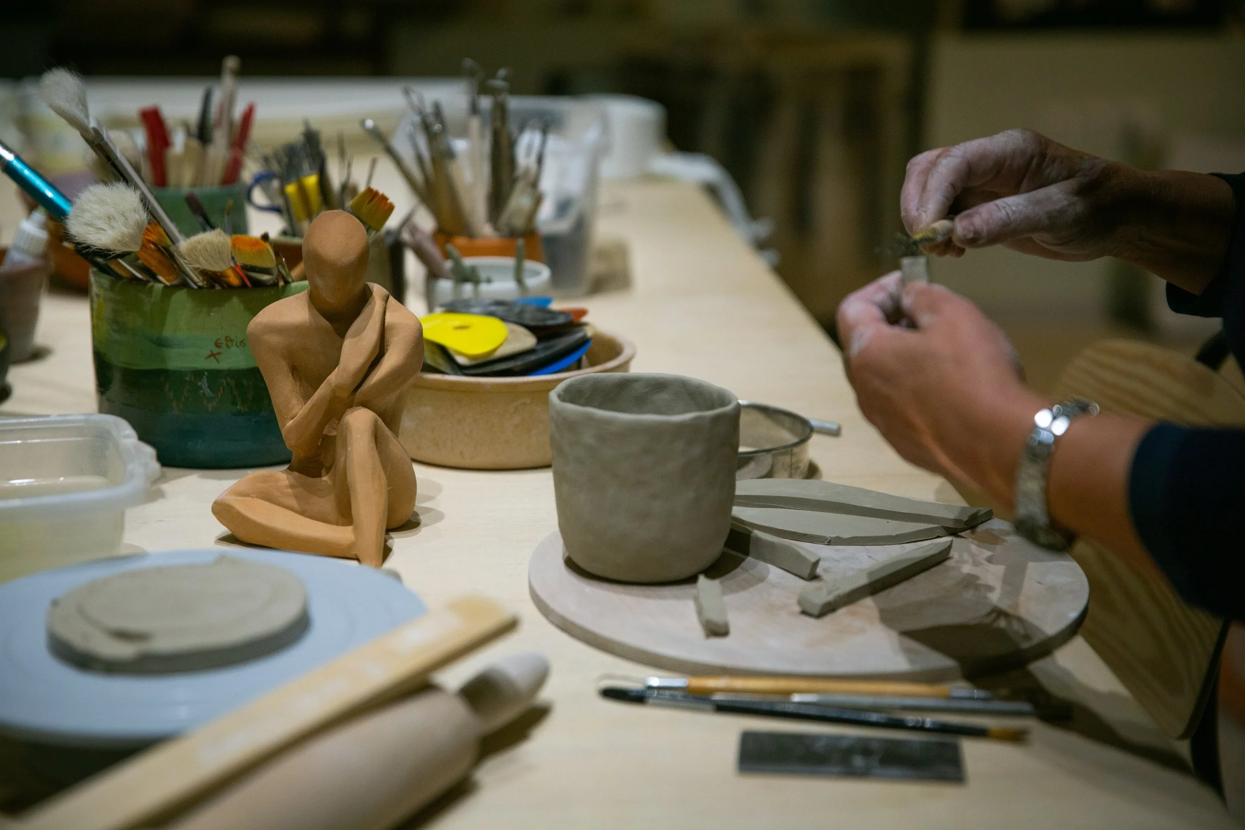A person working on pottery at a craft table, surrounded by various brushes, pottery tools, and ceramic cups, with a small, abstract clay sculpture of a seated figure in the foreground.