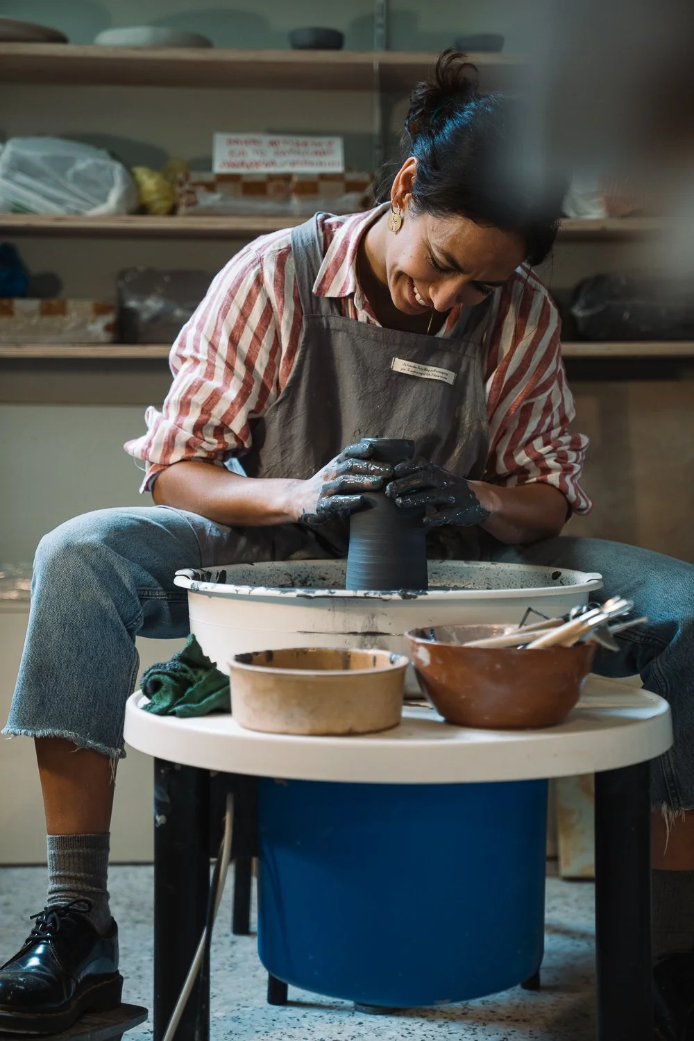 A woman working on a pottery wheel, shaping a piece of black clay in a studio.