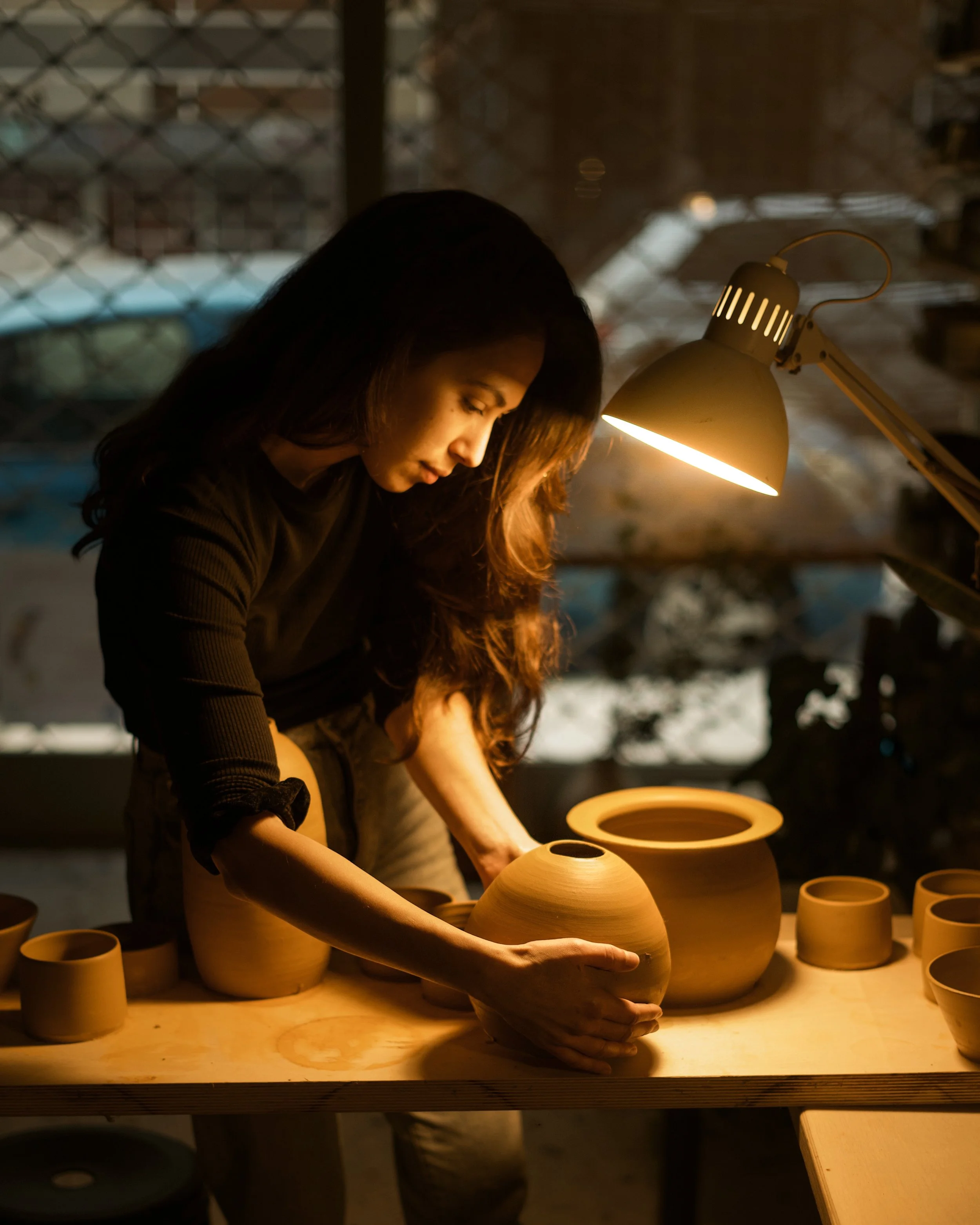 Woman shaping a pottery vase in a workshop with clay pots on a work table illuminated by a desk lamp.