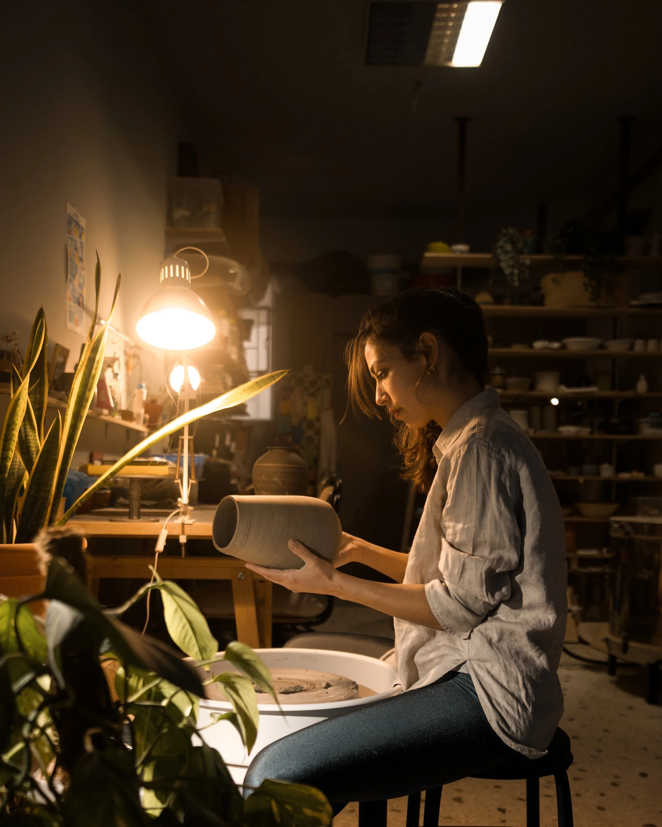 Woman working on pottery in a dimly lit studio with a warm lamp, plants, and shelves filled with pottery and craft supplies.