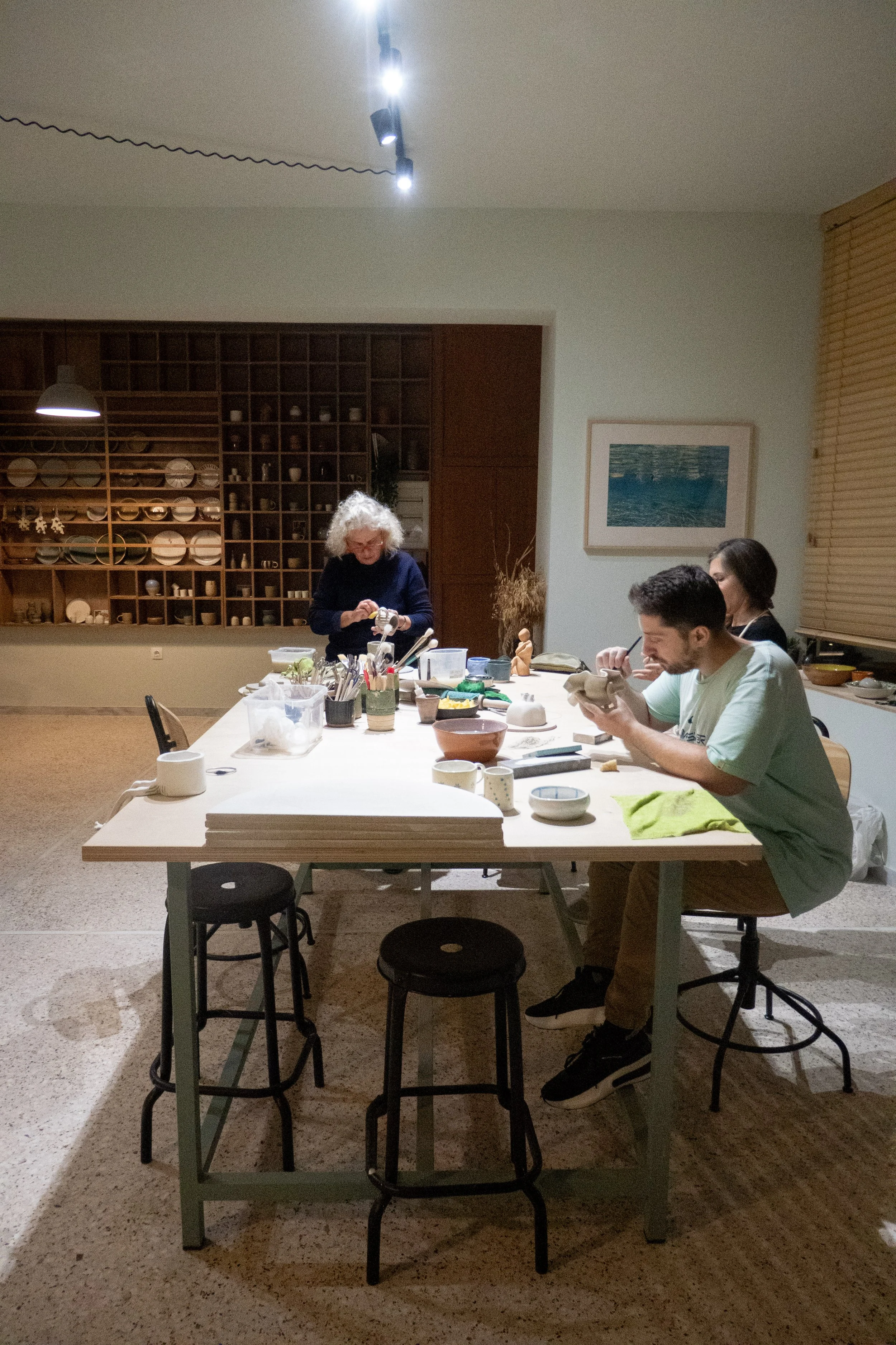 Three people working on a crafting project at a large white table in a well-lit room with wooden shelves and framed picture on the wall.