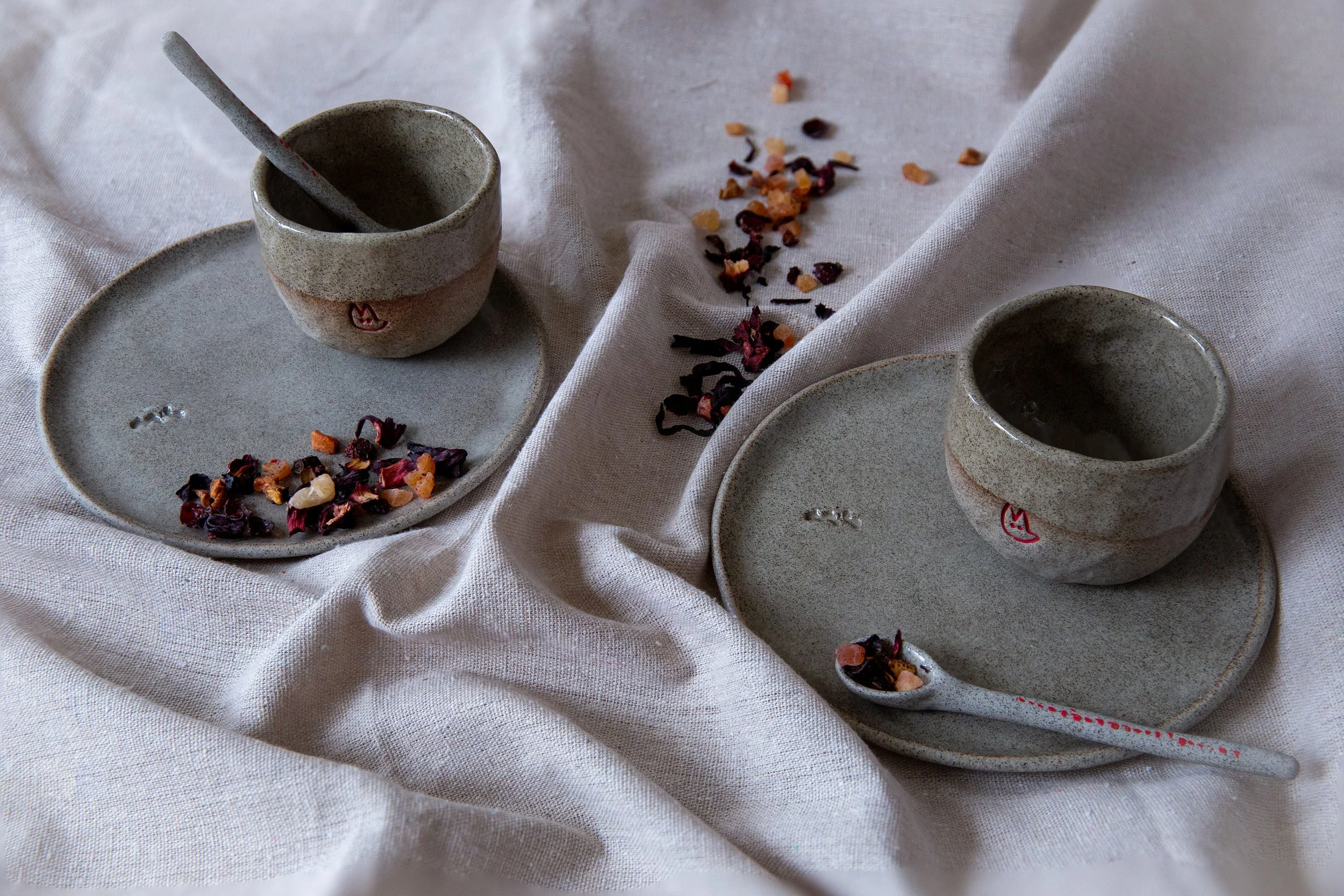 Two ceramic cups with lids on matching plates, with scattered dried rose petals and herbs on a white fabric surface, one cup has a small ceramic spoon with herbs.