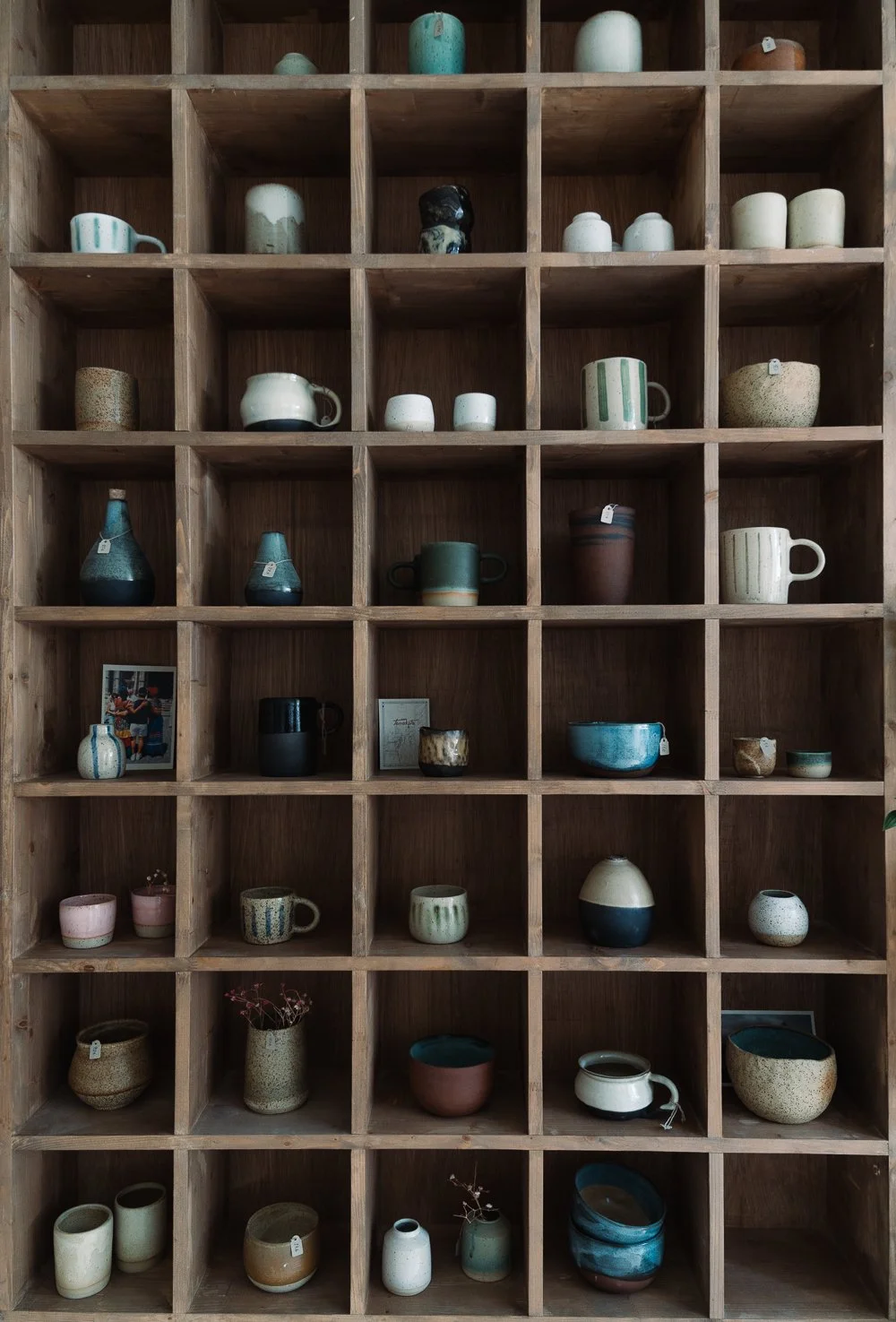 A large wooden display shelf with multiple compartments filled with various ceramic pottery such as mugs, bowls, vases, and cups, some with tags.