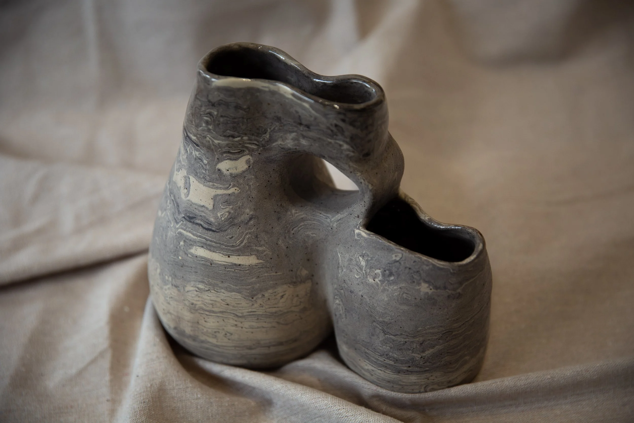 A marble gray and white ceramic vase with a unique shape, featuring two openings and a hole in the middle, placed on a light-colored fabric background.