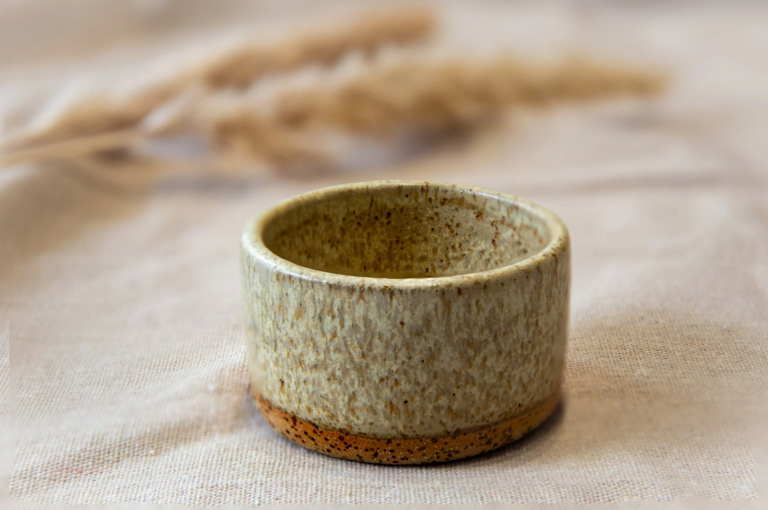 A textured small ceramic cup with earthy tones placed on a light fabric surface, with faint beige woven material in the background.