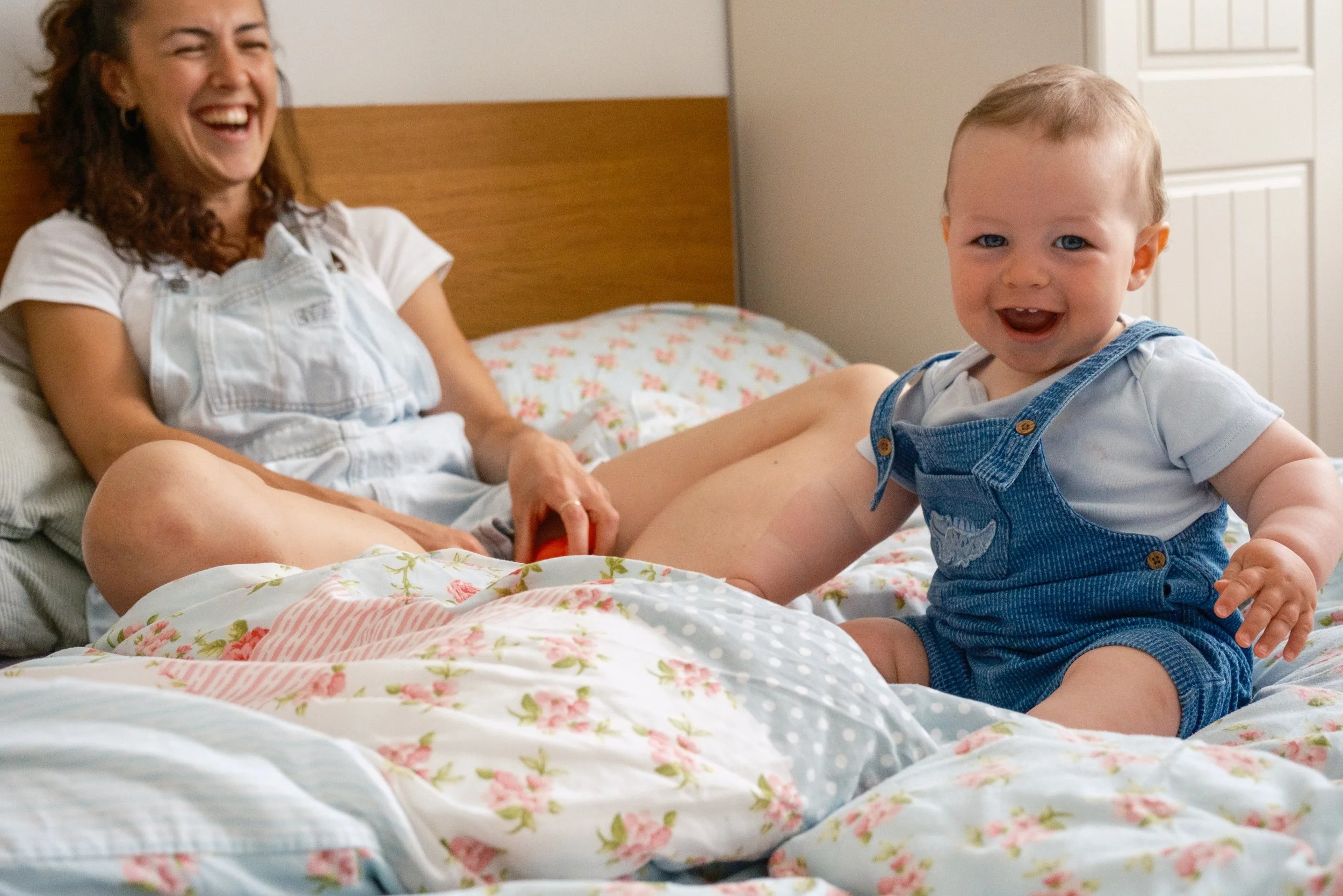 A woman and a baby playing on a bed, both are smiling and laughing, with the woman lying down and the baby sitting up.