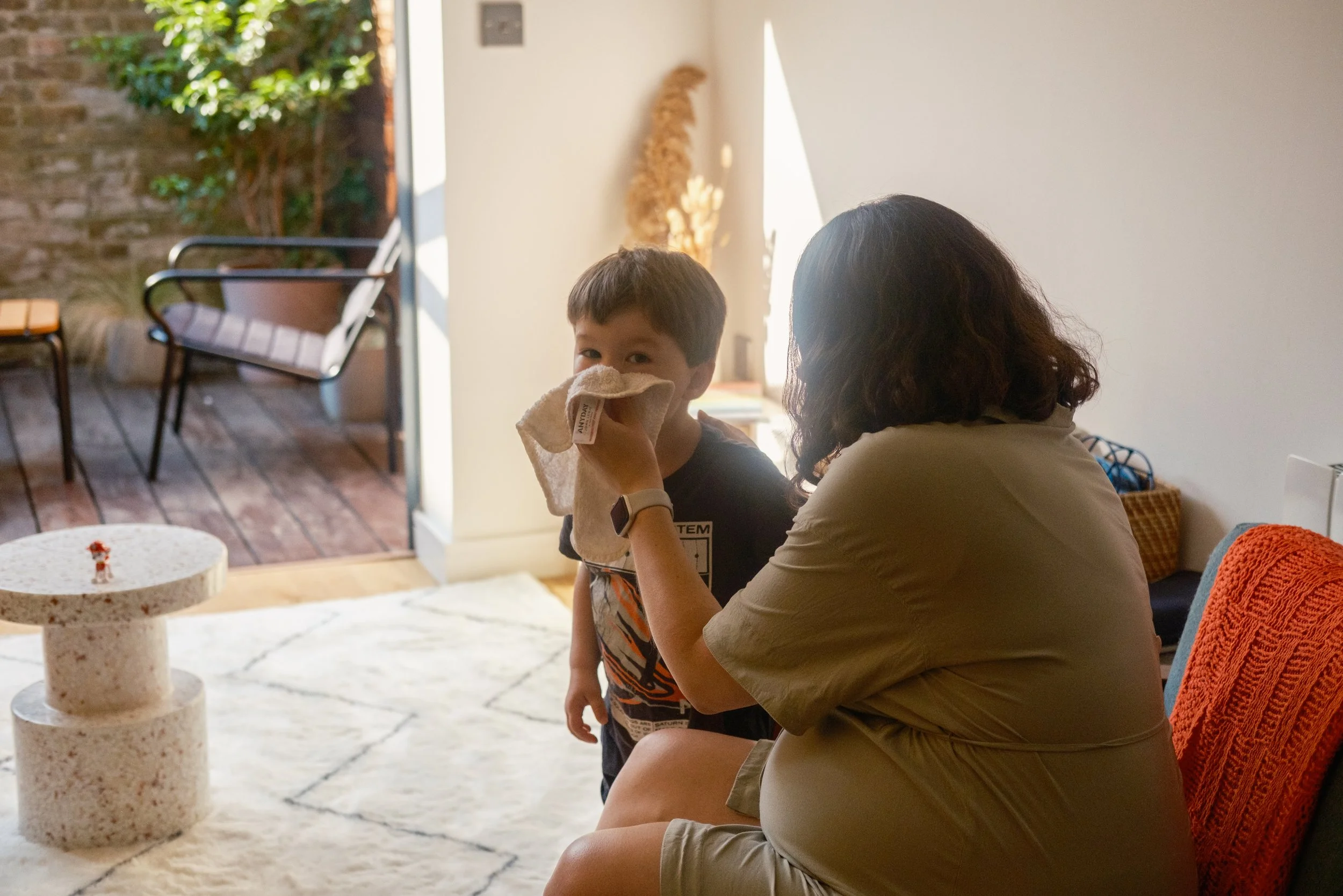 A woman with dark, curly hair sitting on a couch, holding a cloth to a young boy's face as he looks at the camera. The boy is standing near her inside a living room, with an outdoor patio visible through the open door.