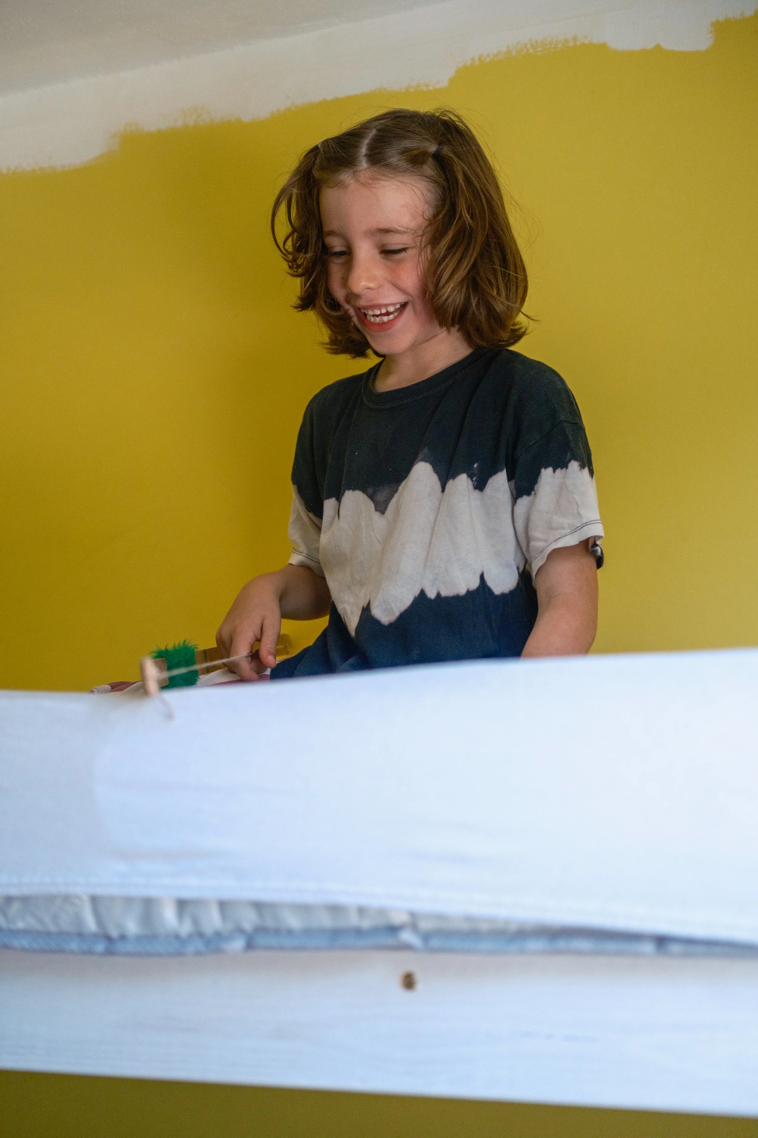 A young girl with shoulder-length brown hair, wearing a black and white tie-dye shirt, laughing while holding a small toy brush with green fur.