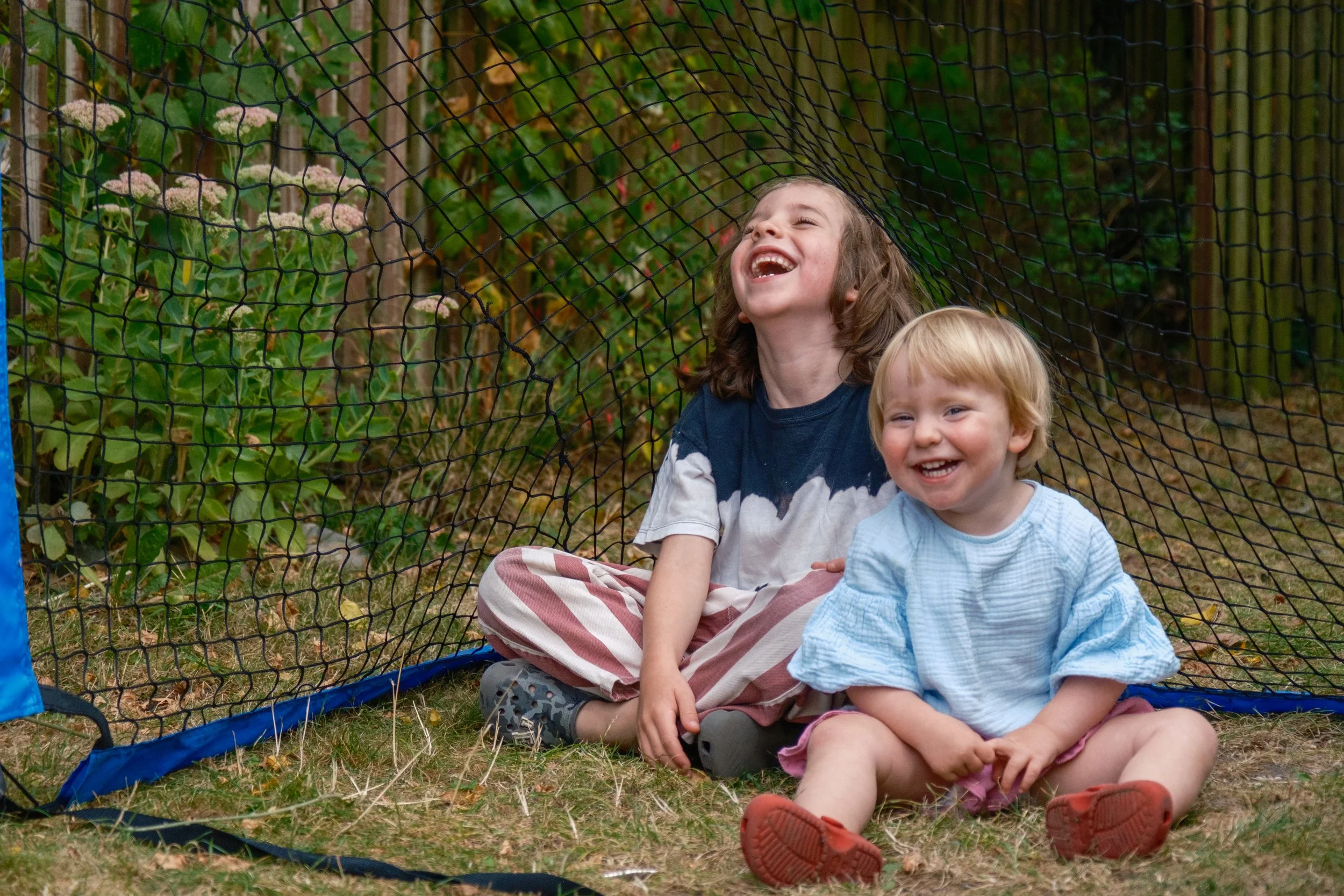 Two children sitting on the grass, laughing under a small outdoor safety net, with bushes and flowers in the background.