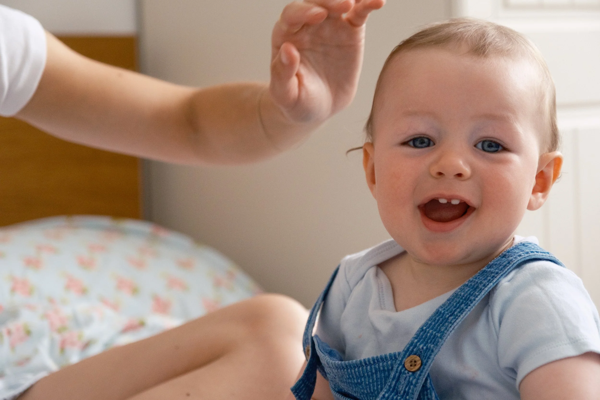 A smiling toddler with blue eyes and light hair, wearing a white shirt and blue overalls, sitting on a bed with floral sheets, in a room with sunlight coming through a window.