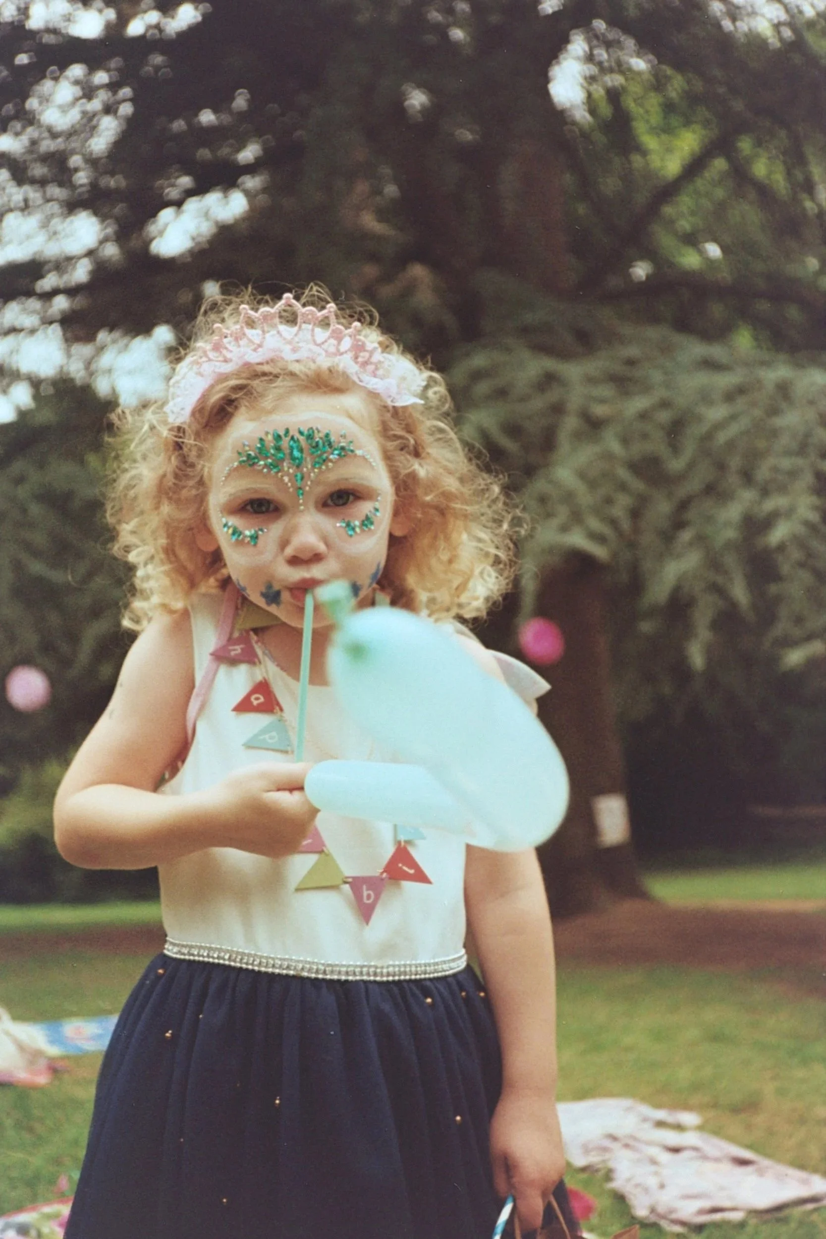 A young girl with curly red hair and decorated face, wearing a pink tiara and a necklace made of colorful triangles, holding a blue balloon shaped like a bunny, standing outdoors with trees in the background.