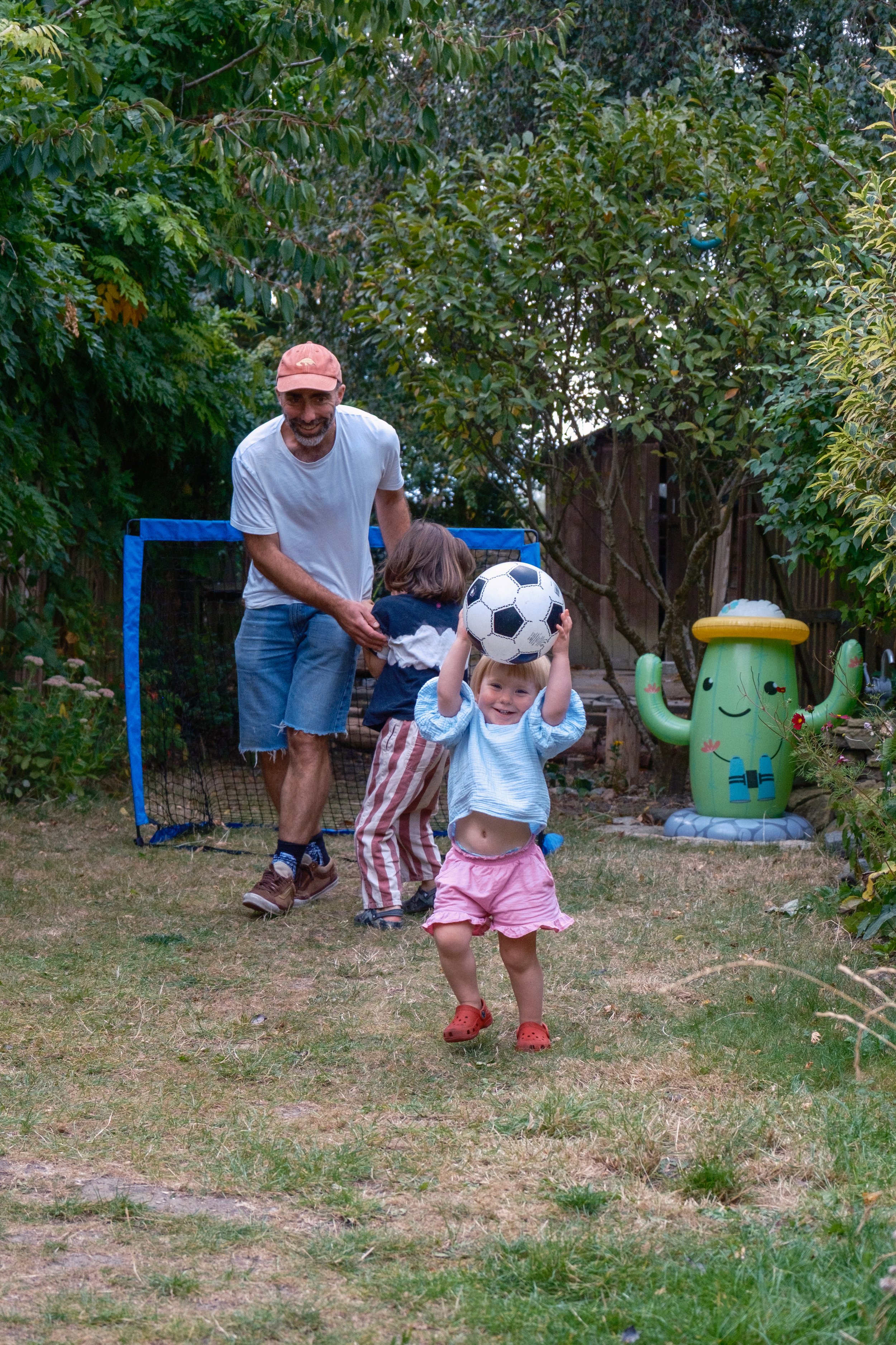 A man and two children playing with a soccer ball in a backyard. The girl in the foreground is holding the ball above her head, smiling. The boy and man are in the background, near a small goal net. There are trees and garden decorations around.