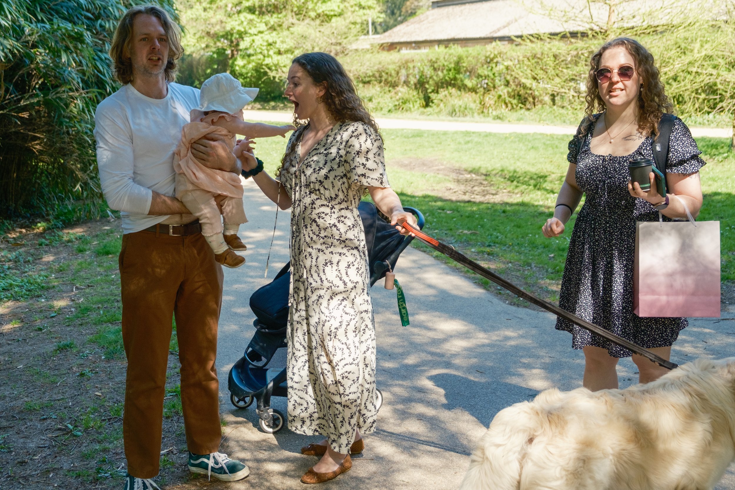 A group of four people outdoors on a sunny day, with a man holding a baby, a woman with a stroller, another woman holding a coffee cup and shopping bag, and a large dog partially in the foreground.