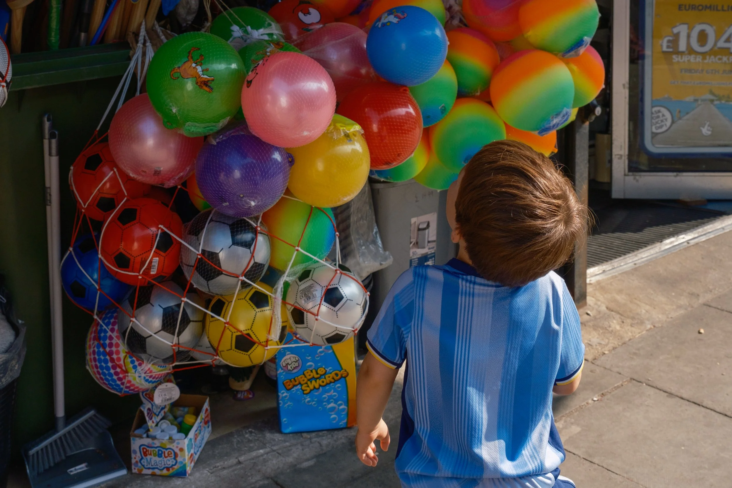 A young boy with brown hair wearing a blue striped shirt stands near a display of colorful plastic balls and toy soccer balls outside a store.