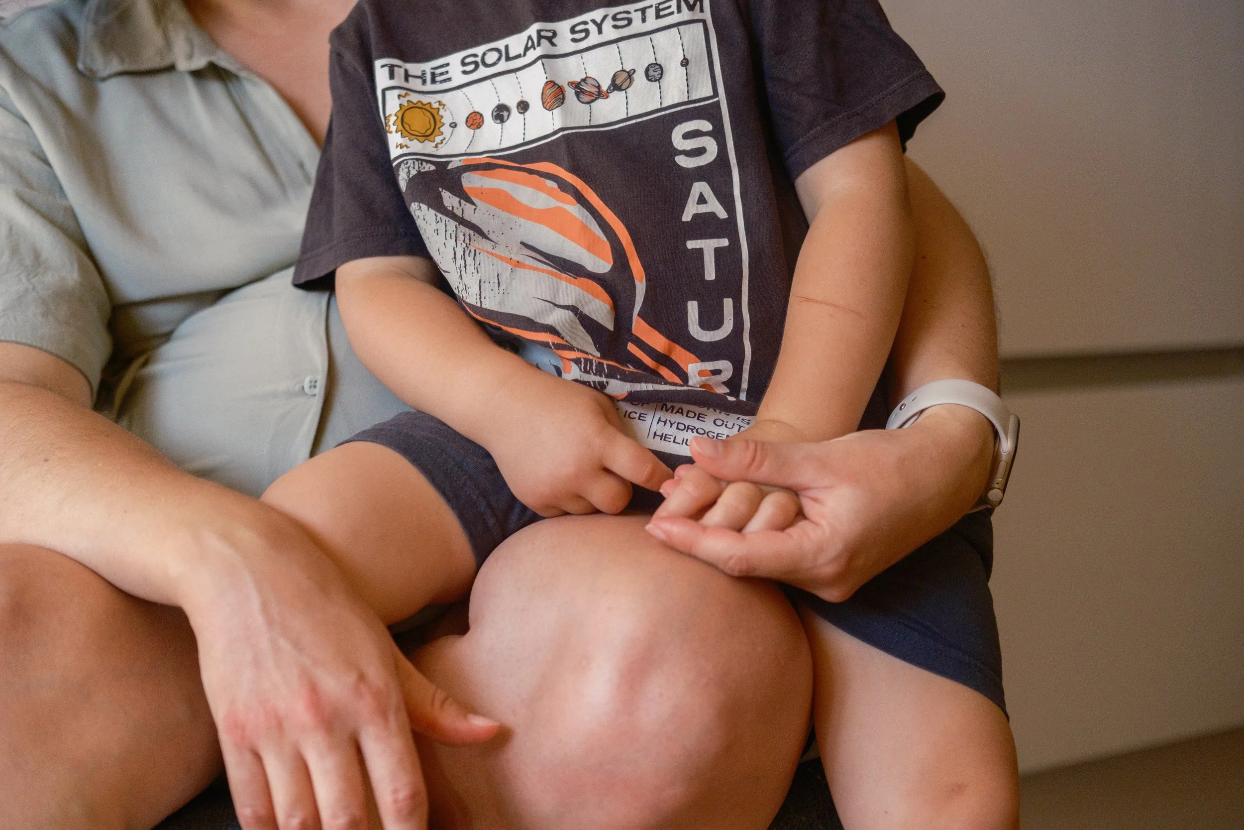 A child sitting on an adult's lap, holding the adult's hand, with the adult's arms visible, wearing a beige shirt and a digital wristwatch, while the child wears a dark T-shirt with space-themed graphics and text.