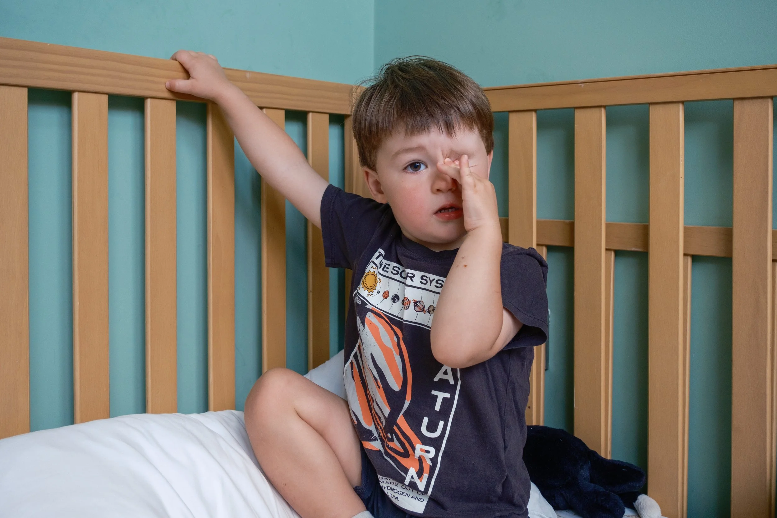 Young boy sitting on a bed with a wooden headboard, holding a finger to his nose and looking surprised or worried.