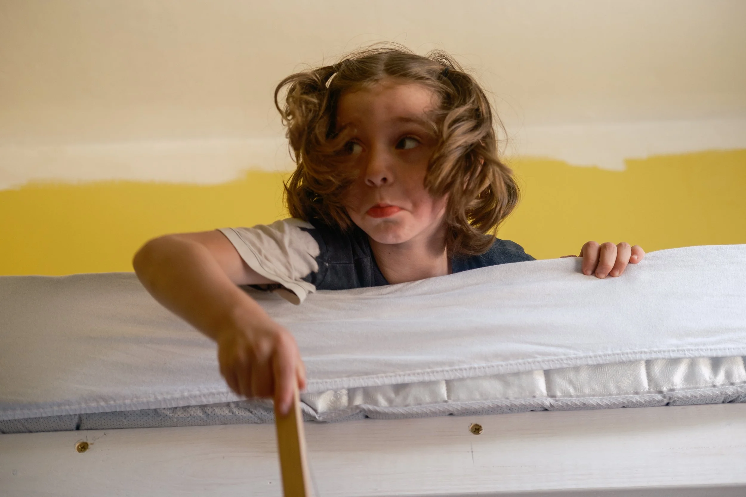 A young child with curly hair peeks over the edge of a bed, looking to the side with a thoughtful expression.