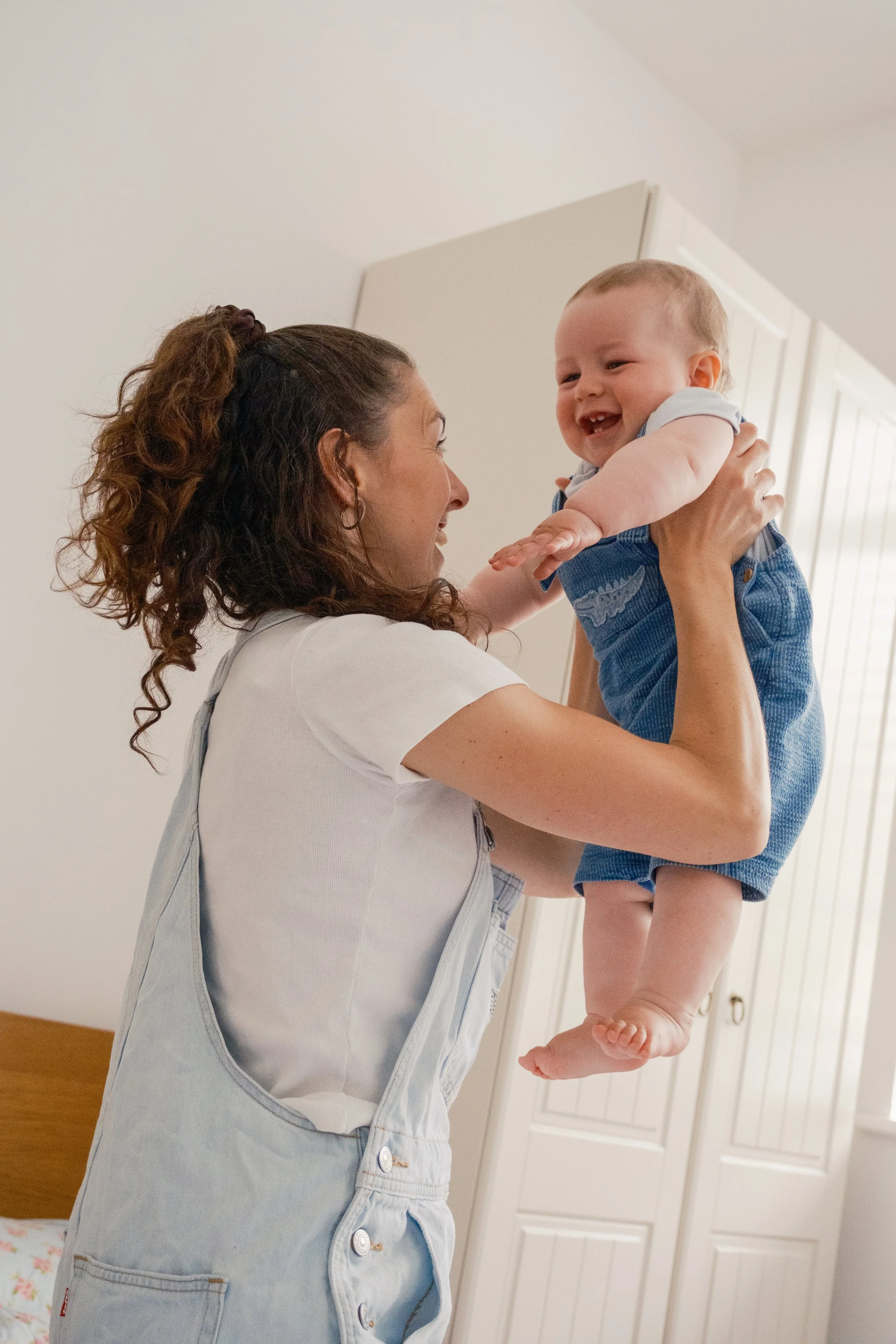 A woman lifting a laughing baby in a bedroom with white walls and a wooden bed frame.