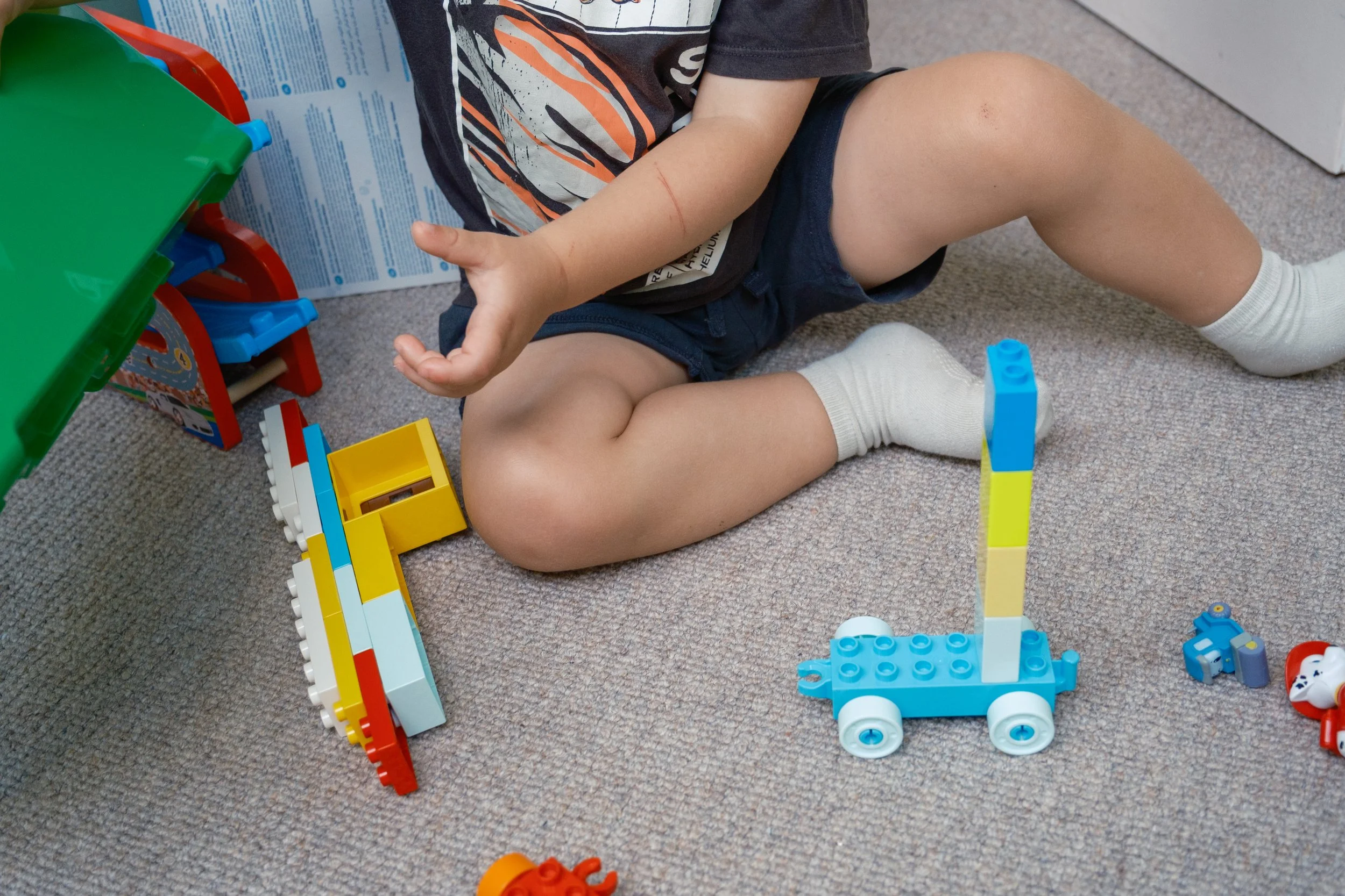 Child playing with LEGO bricks on a carpeted floor, with a green table and a makeshift fort made from a cardboard box in the background.