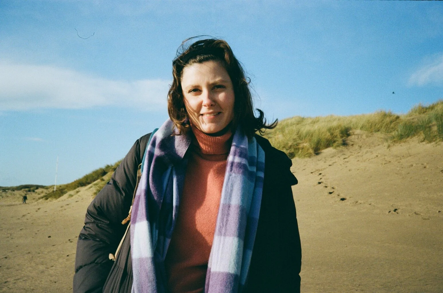 Woman standing on sand dune beach with grassy top and blue sky, wearing a black jacket, pink turtleneck, and purple checkered scarf.
