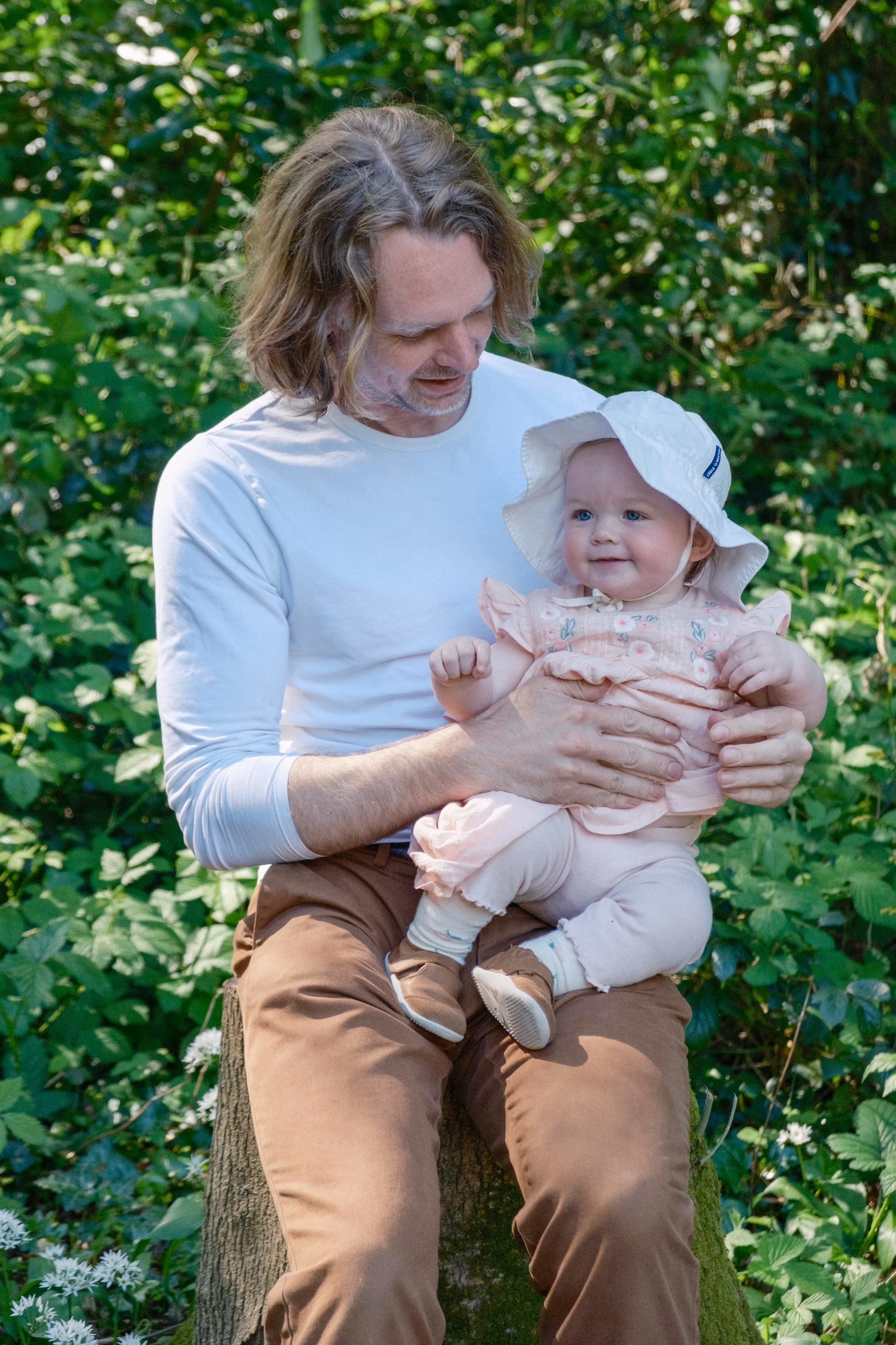 A man with shoulder-length hair holding a smiling baby girl wearing a sun hat outdoors surrounded by greenery.