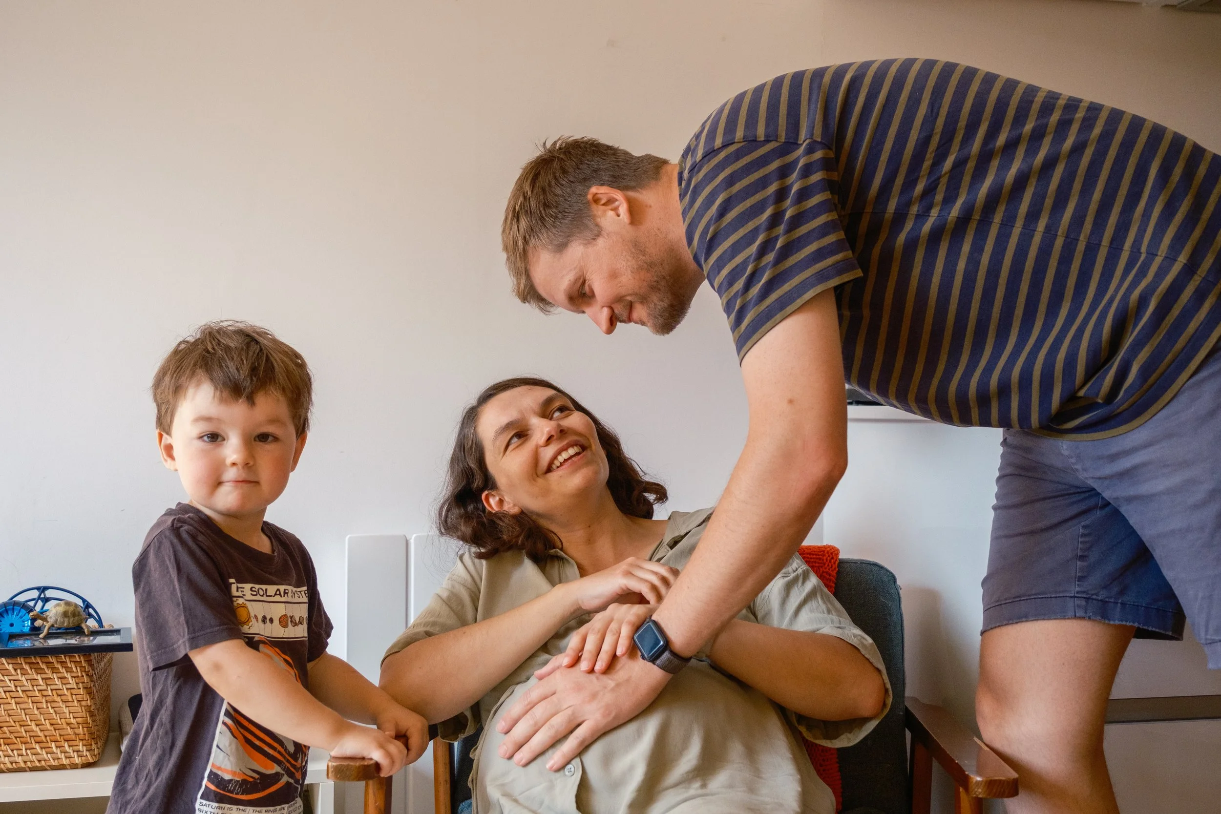 A woman sitting in a chair with a baby bump, smiling and looking up at a man leaning over her, holding her hand. Two young boys, one standing and one sitting, are nearby. The scene appears warm and affectionate, suggestive of a family moment.
