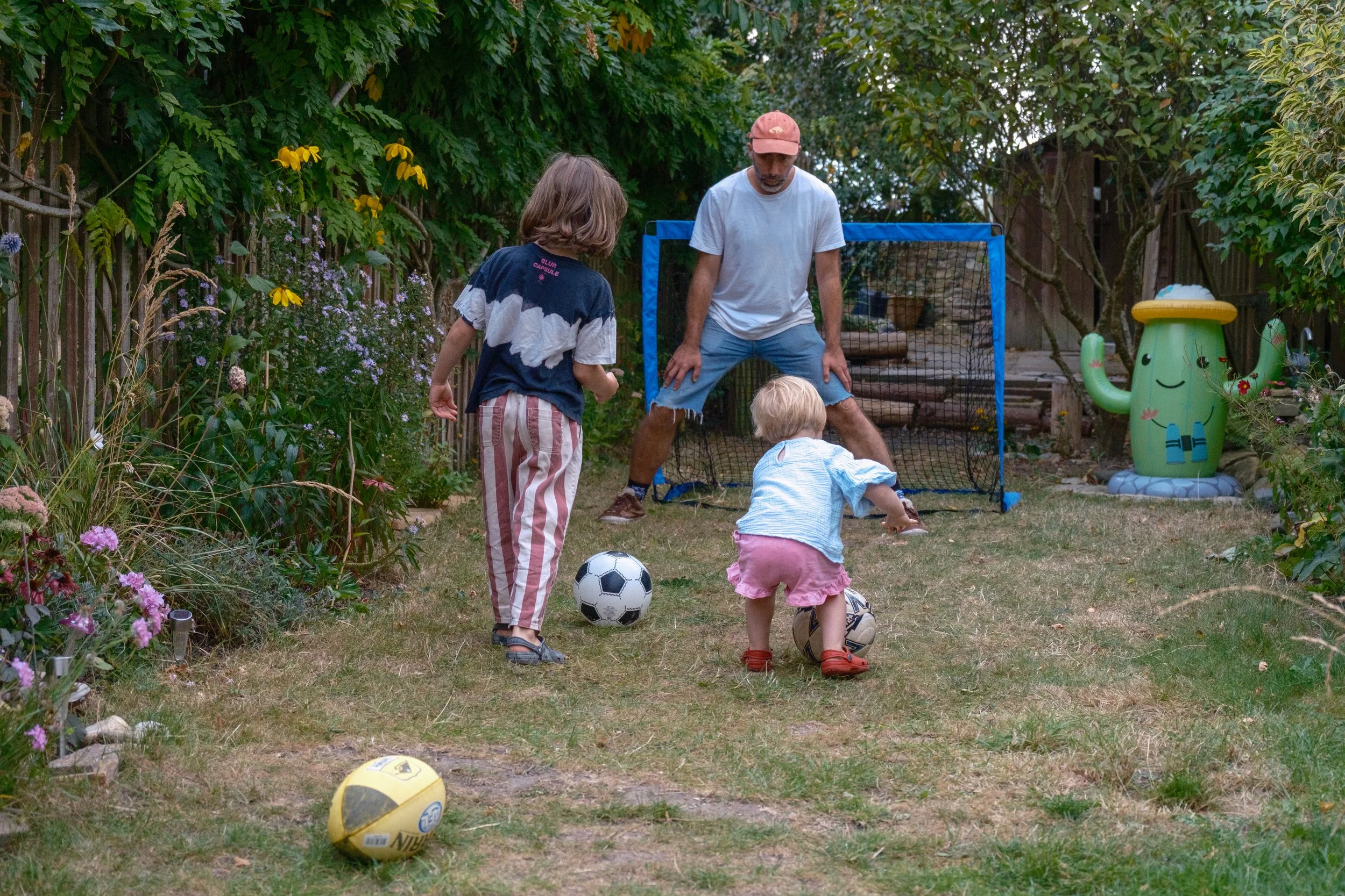 A man and two young children playing soccer in a backyard garden with trees, flowers, and garden decorations.