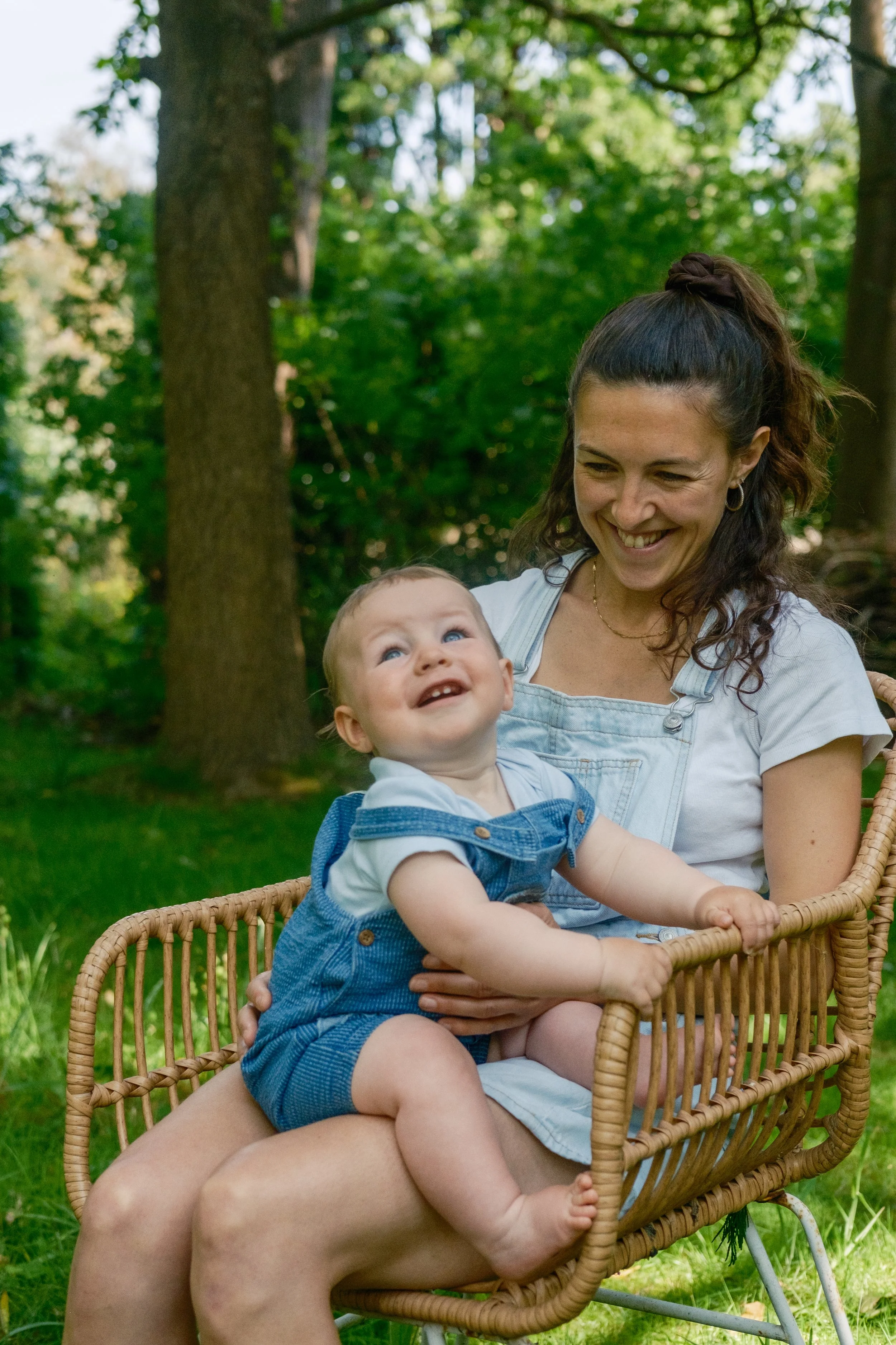 A woman and a young child sitting on a wicker bench outdoors in a green, wooded area, smiling and looking at each other.