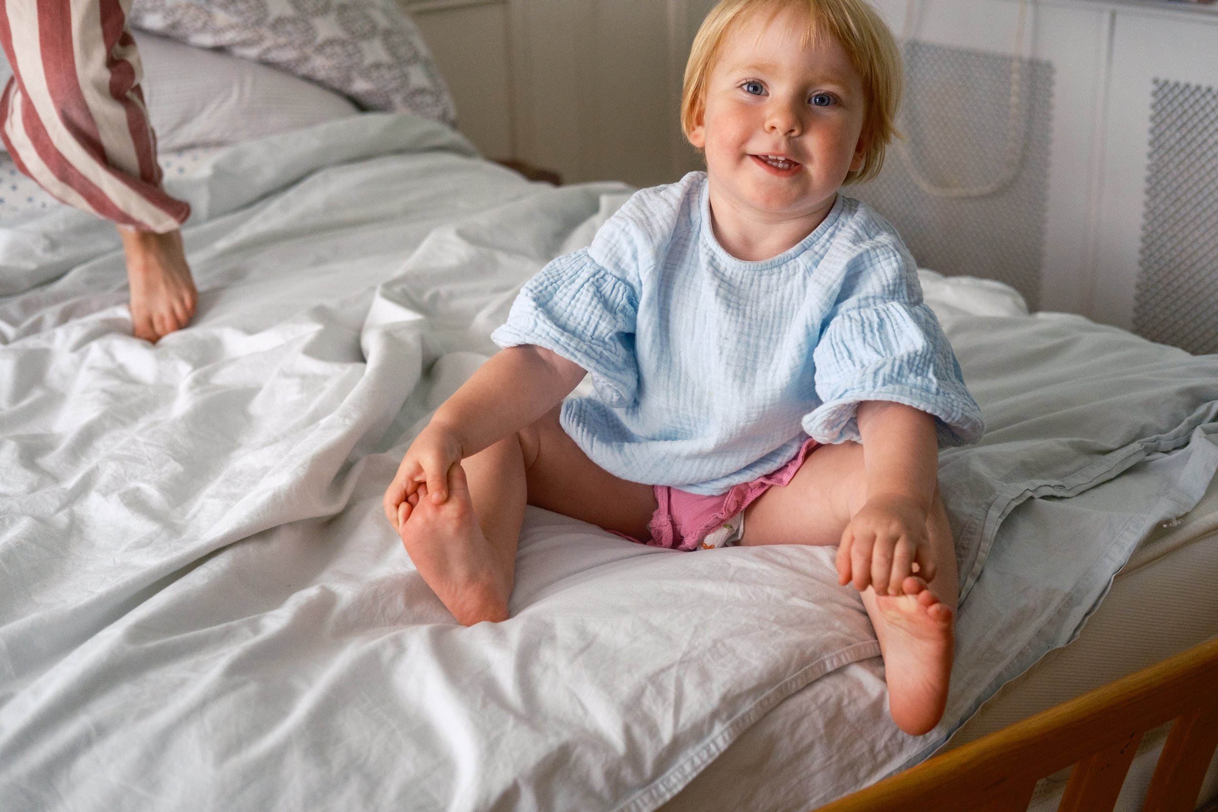 A young child with blonde hair, wearing a light blue top and pink diaper, sitting on a white bed with a happy expression.