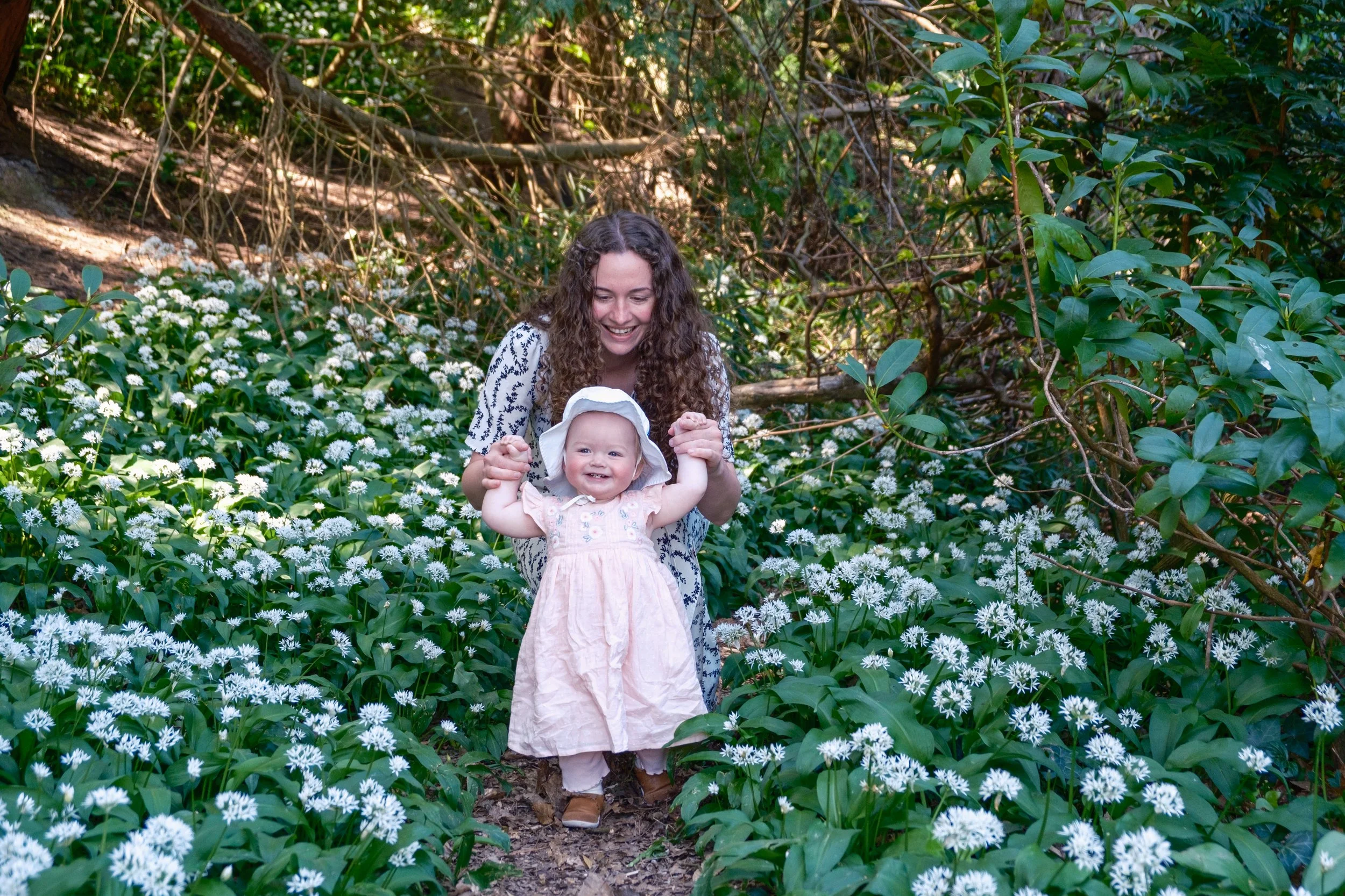 A woman with curly brown hair smiling and holding hands with a young girl in a pink dress and sun hat, walking through a forested area with white flowers on the ground.