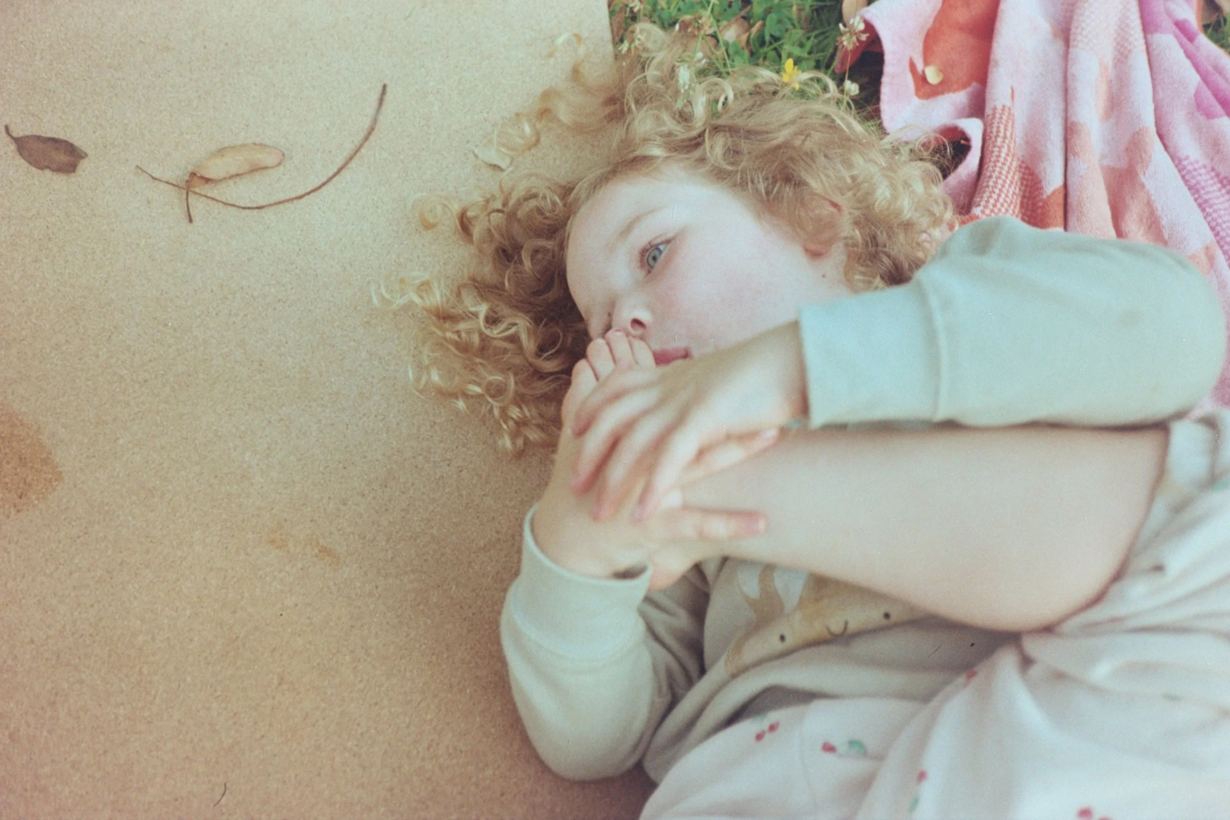 A young girl with curly blonde hair lying on the ground, resting her head on a towel, appearing thoughtful or relaxed.
