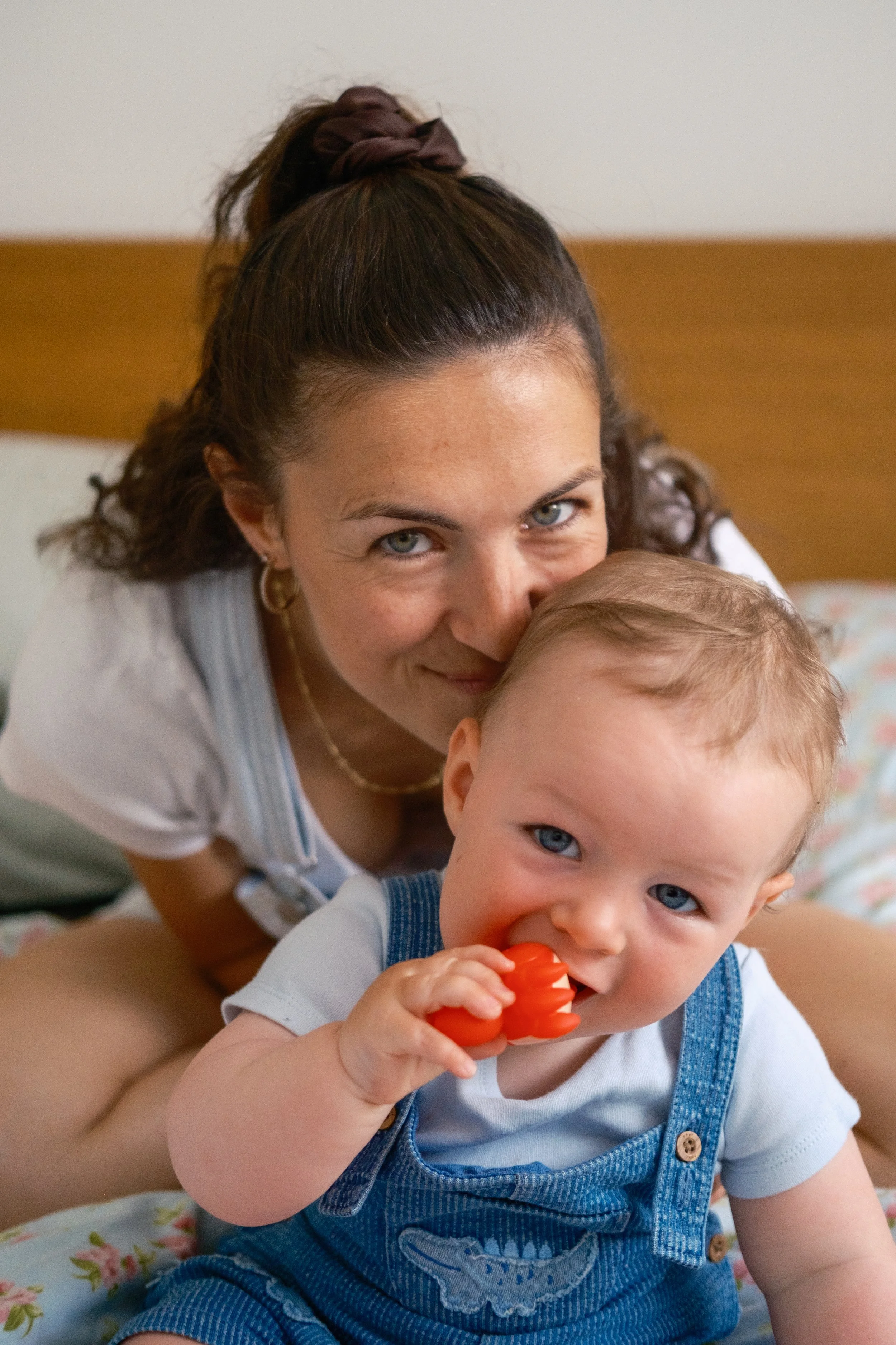 A woman and a young boy with blue eyes, close together on a bed, smiling at the camera. The boy is holding a red toy and has light brown hair, wearing overalls and a light-colored shirt.