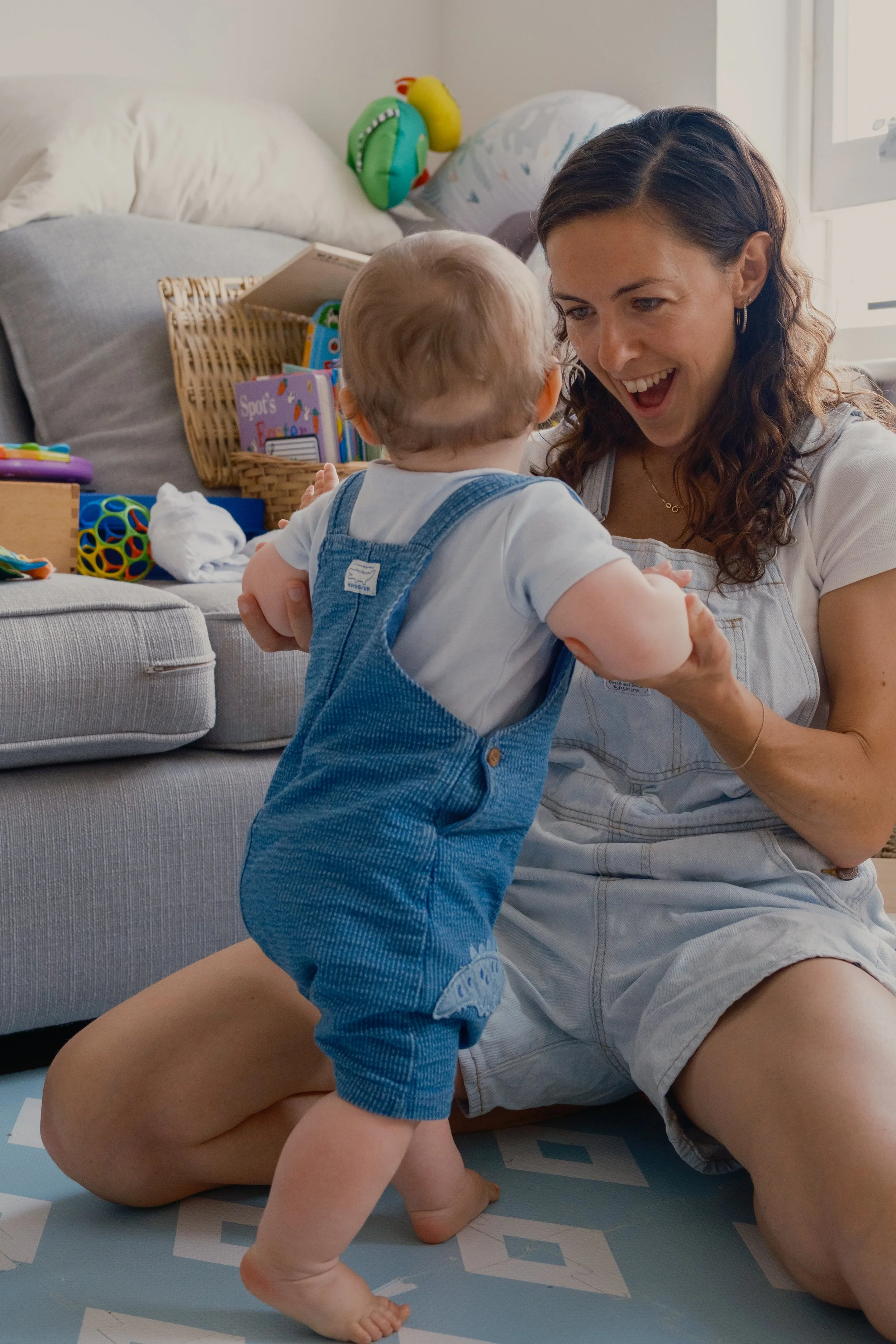 A woman and a toddler playing together on a patterned mat in a living room filled with toys; the woman is smiling and holding the child's hands as he climbs.