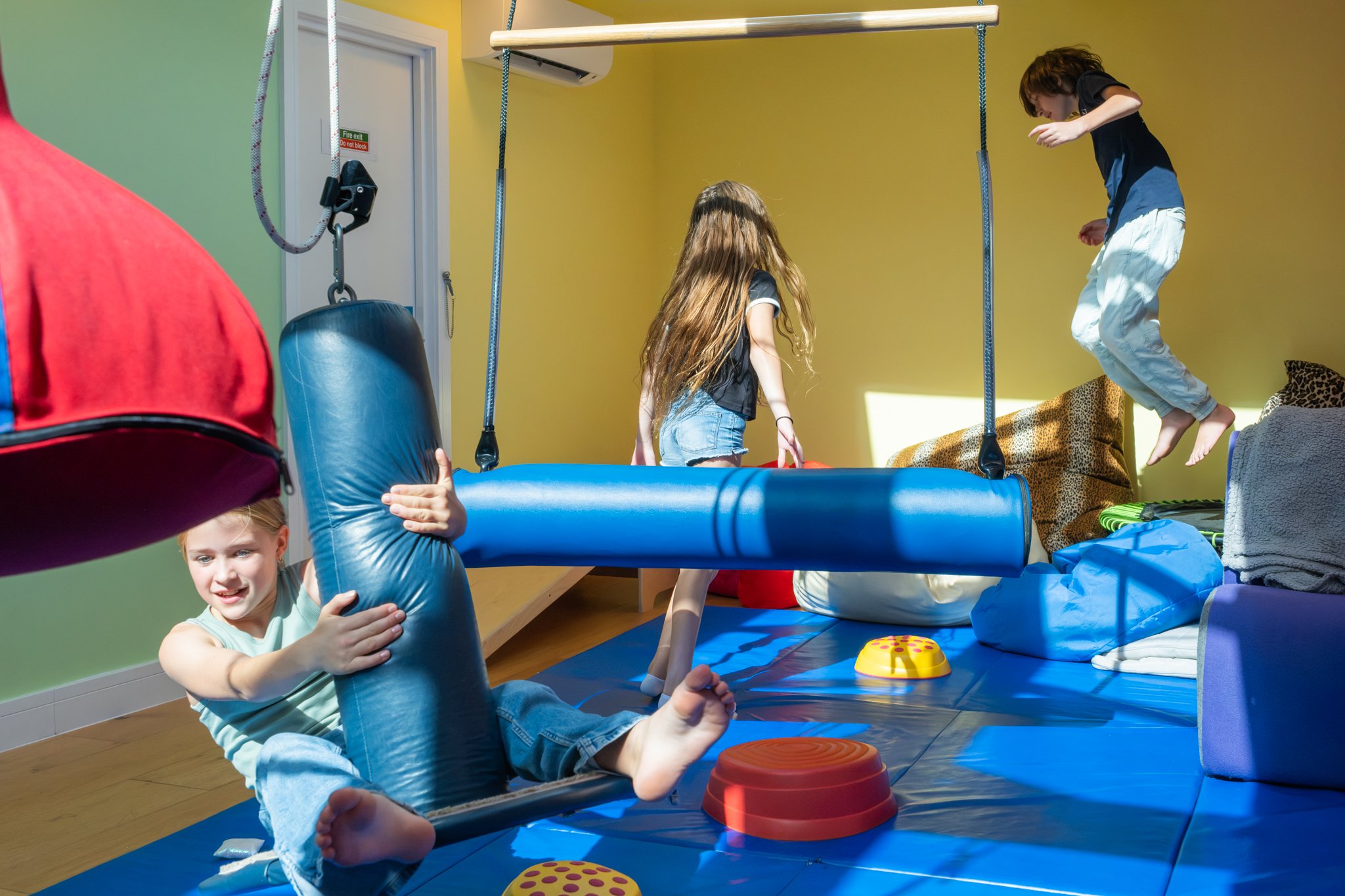 Children playing on a blue padded indoor trampoline, jumping and climbing, with various soft play equipment around them, in a brightly decorated playroom with yellow and green walls.