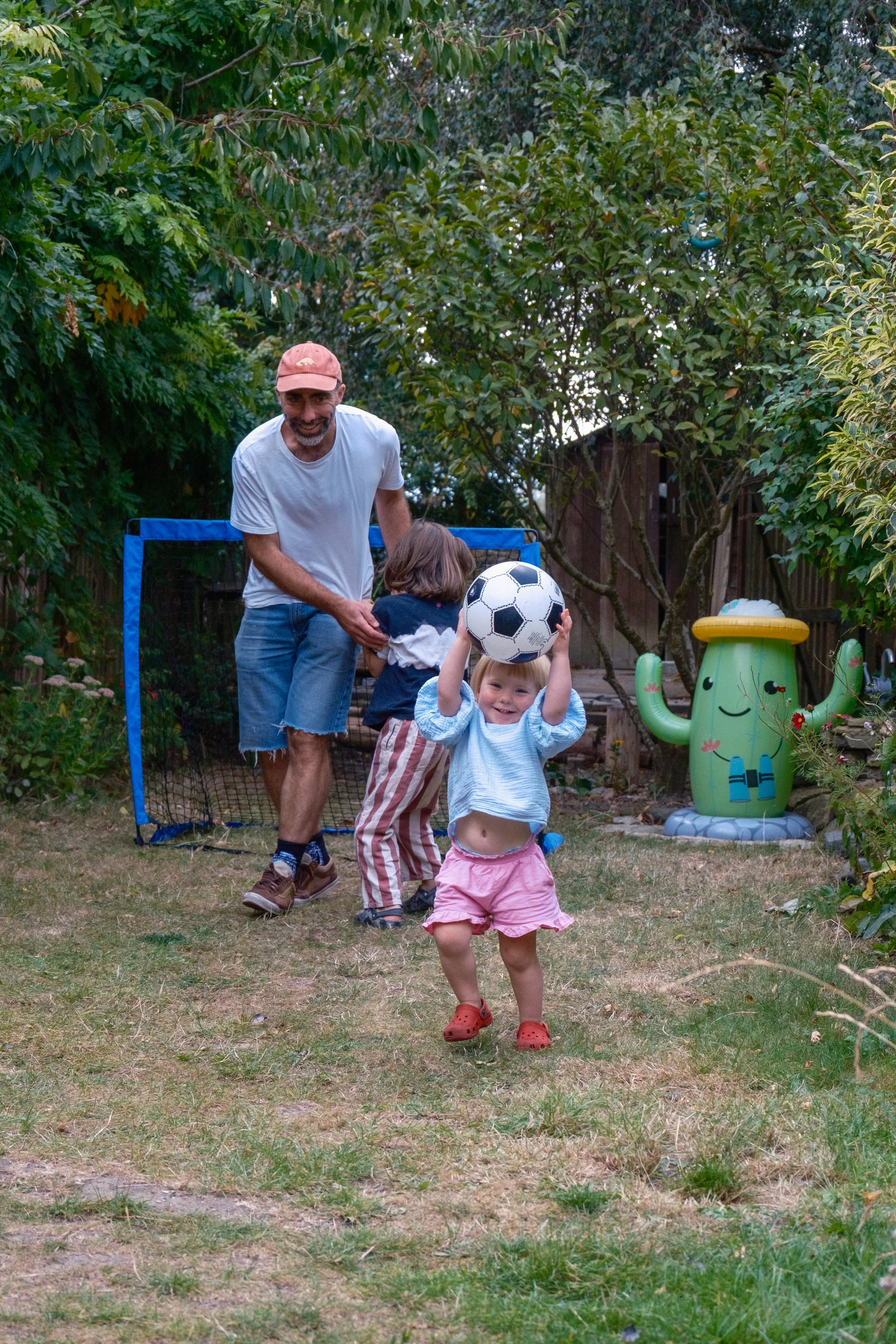 Family playing soccer in backyard with trees and a fun cactus-shaped decoration.