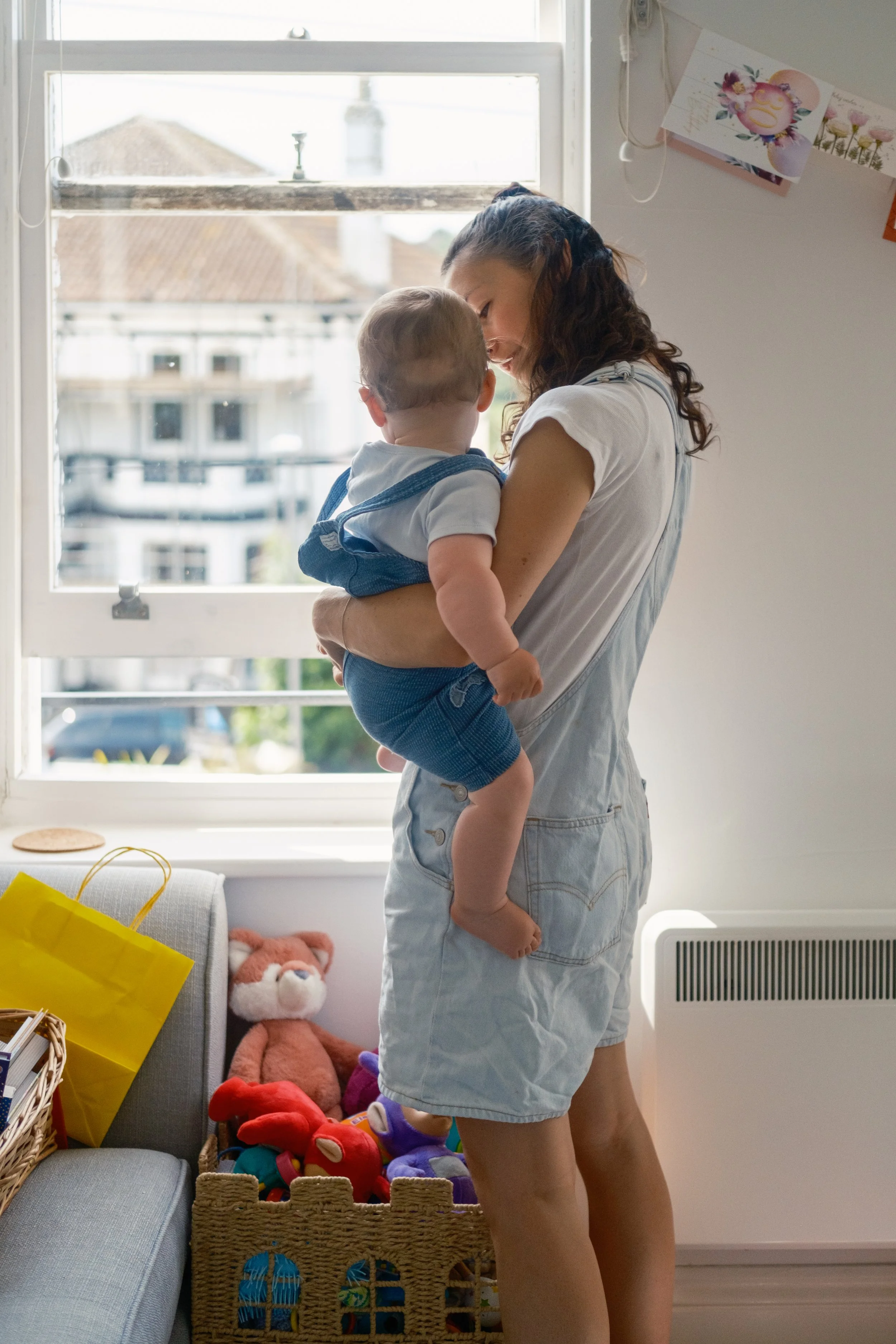 A woman holding a young child near a window in a room decorated with toys and cards.